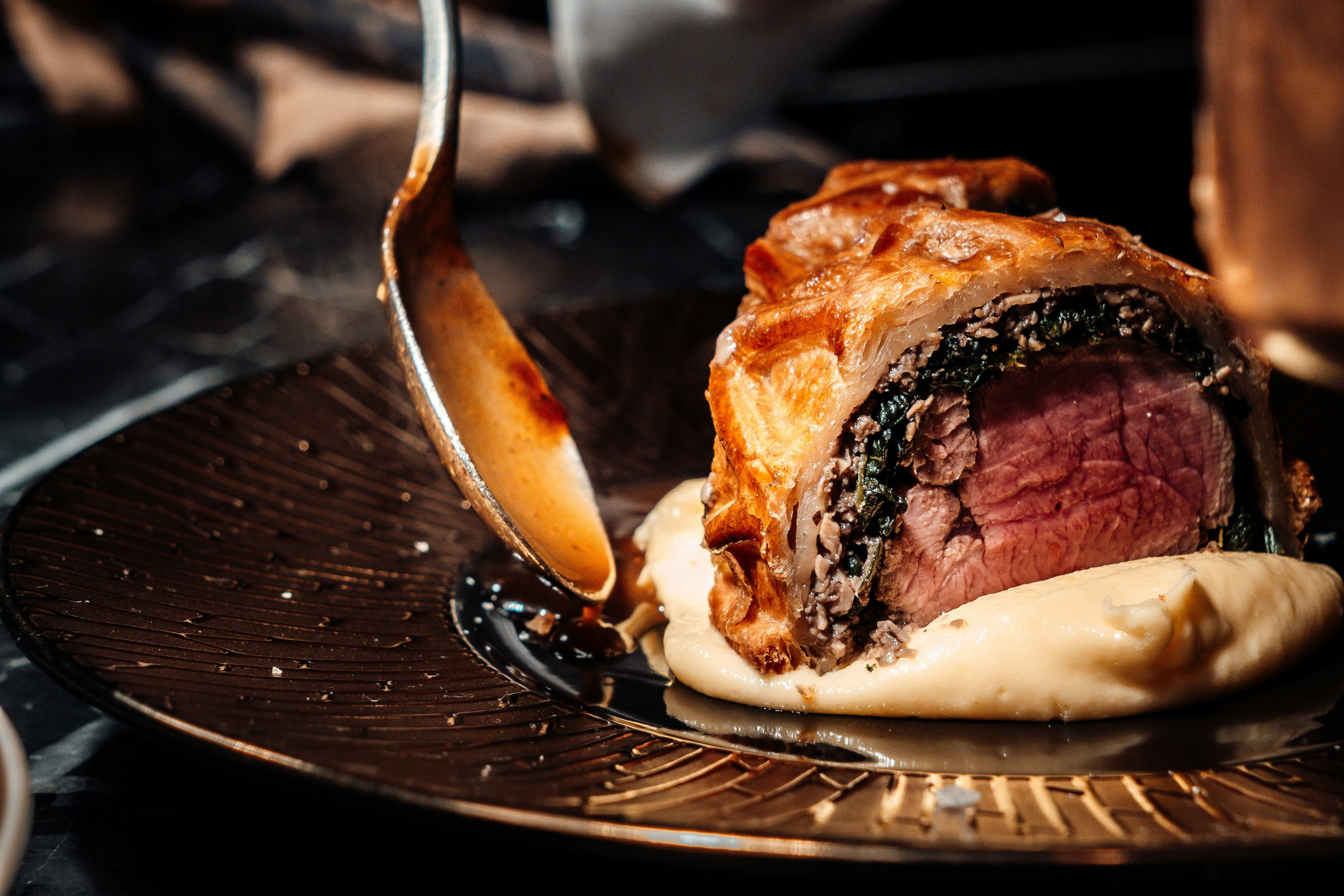 A beef Wellington with a pink center, wrapped in biscuit dough, sliced open to show a layer of spinach and minced meat, served on a bed of creamy mashed potatoes on a decorative black plate.