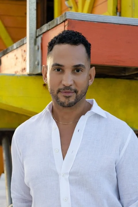 A man with short dark curly hair, trimmed beard, wearing a white button-up shirt standing outdoors in front of colorful wooden structures.