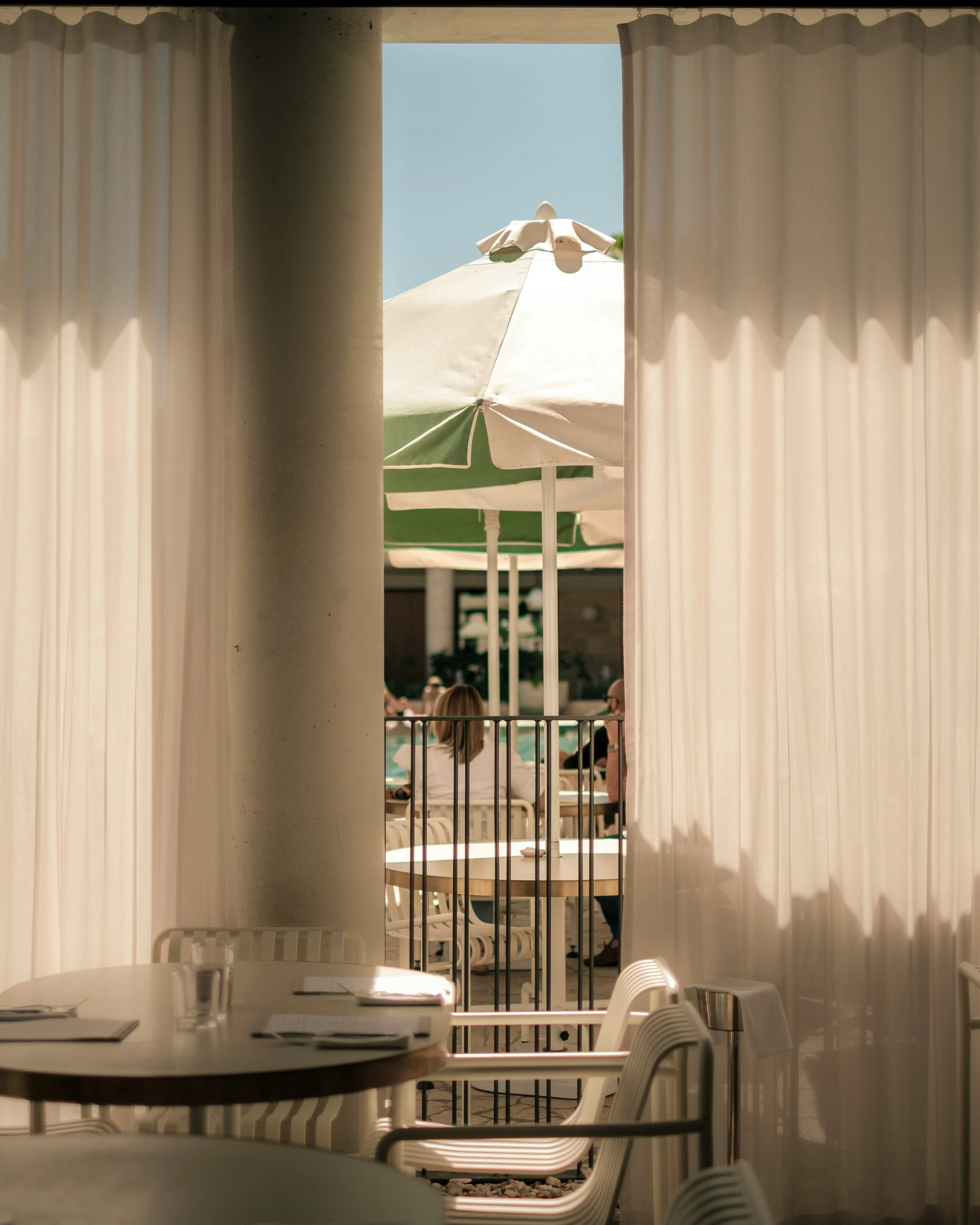 Interior of a restaurant with beige curtains and tables set with menus and glasses, view outside showing a patio with white umbrellas and people sitting by the pool.