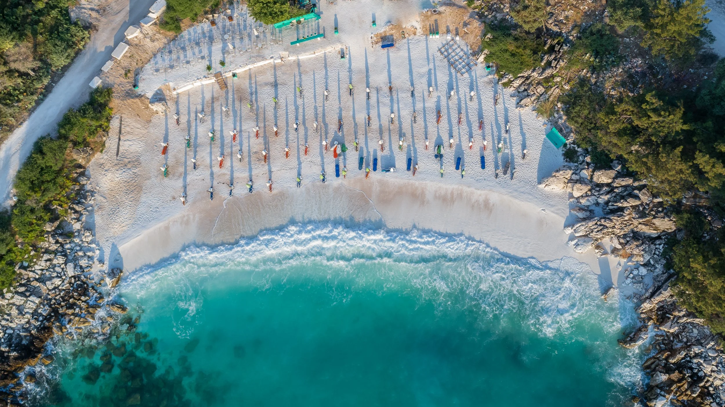 Aerial view of a beach with colorful umbrellas and lounger chairs on the sand, near turquoise water, surrounded by green trees and rocks.