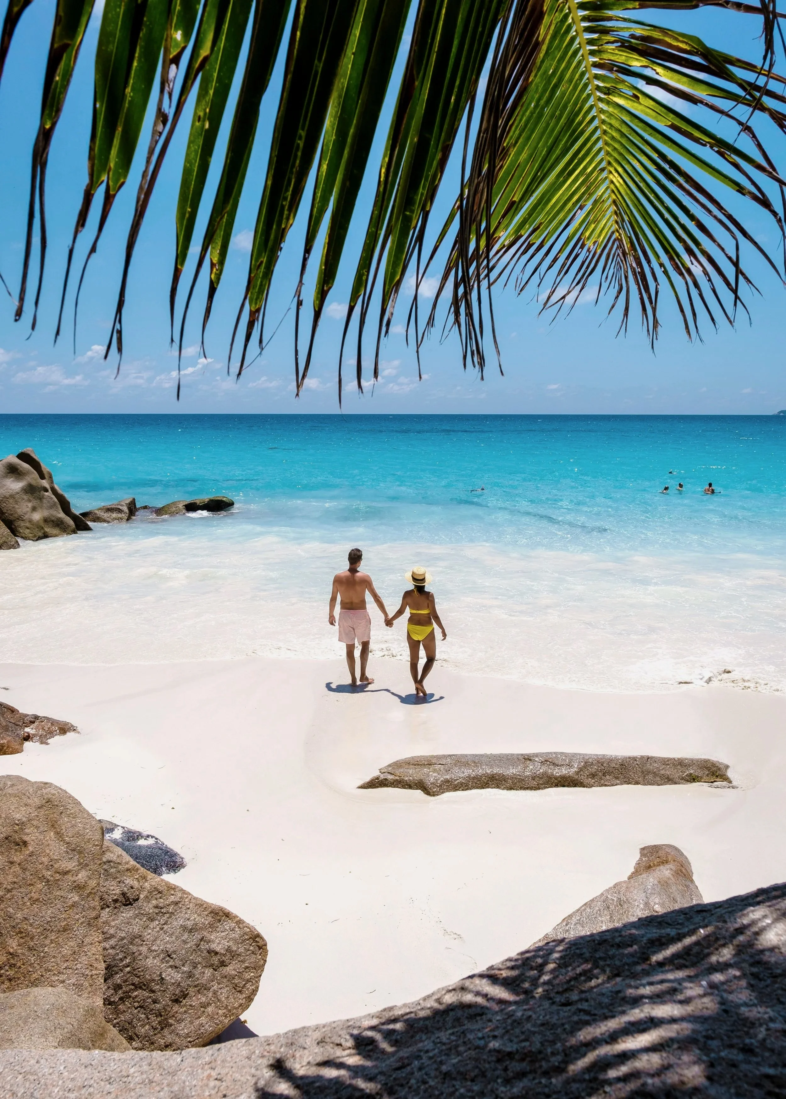 Couple holding hands walking on a sandy beach with turquoise water, rocks, and palm leaves overhead.