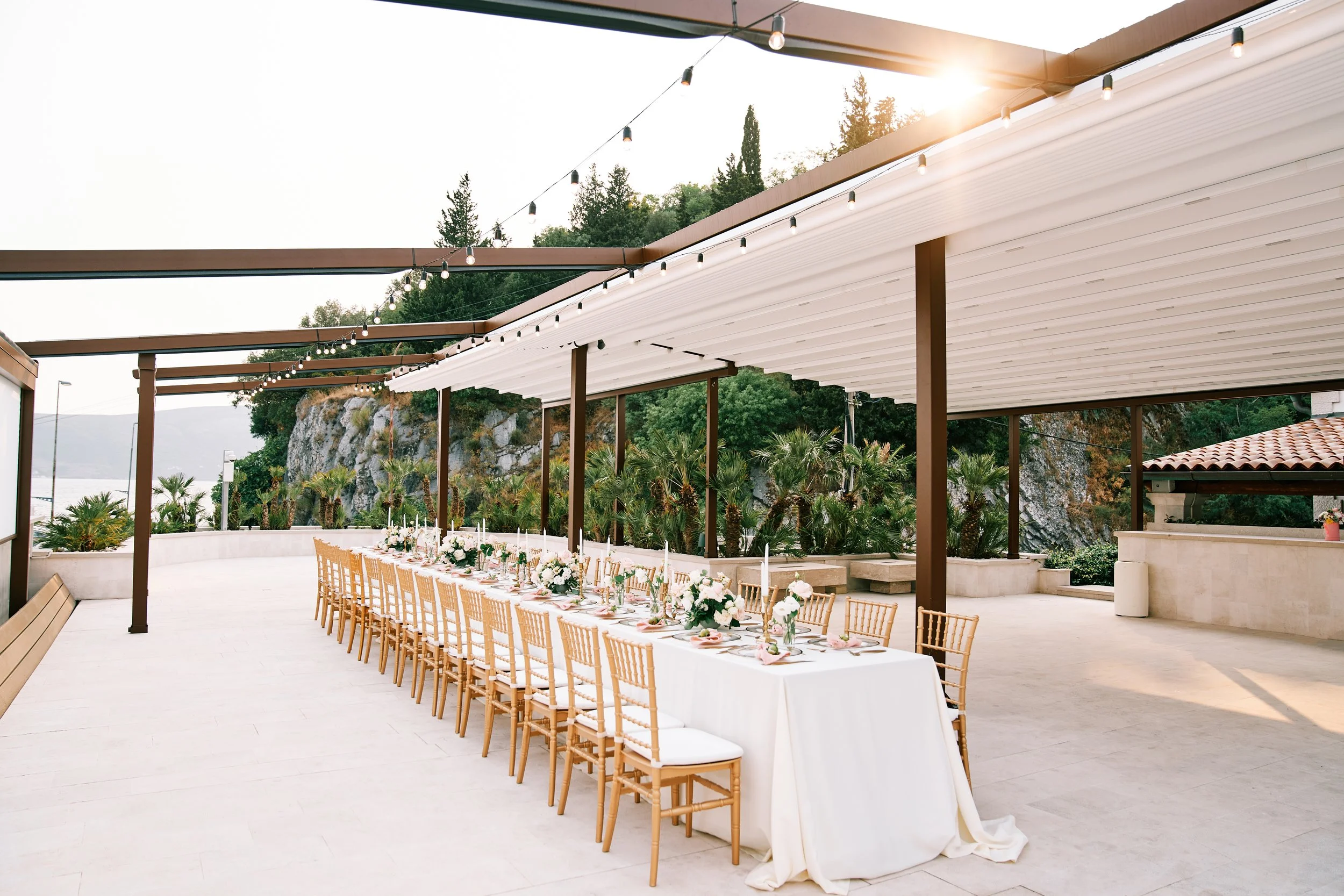 Outdoor event space with a long dinner table decorated with flowers, candles, and place settings, under a white pergola with string lights, surrounded by greenery and mountains.