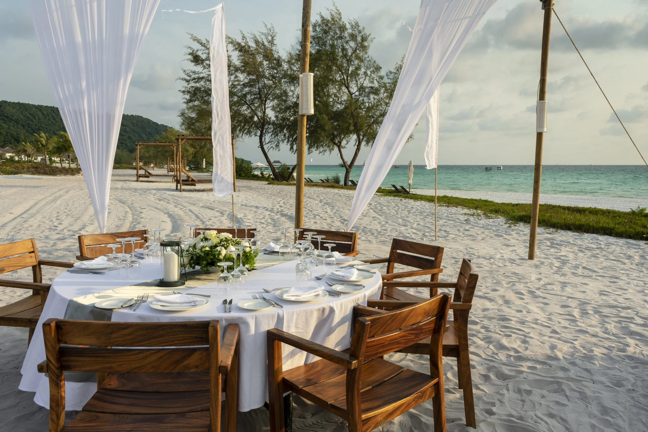 A table set for a beachside meal with white tablecloth, plates, glasses, cutlery, and floral centerpiece, under a canopy with white drapes on a sandy beach near the ocean.