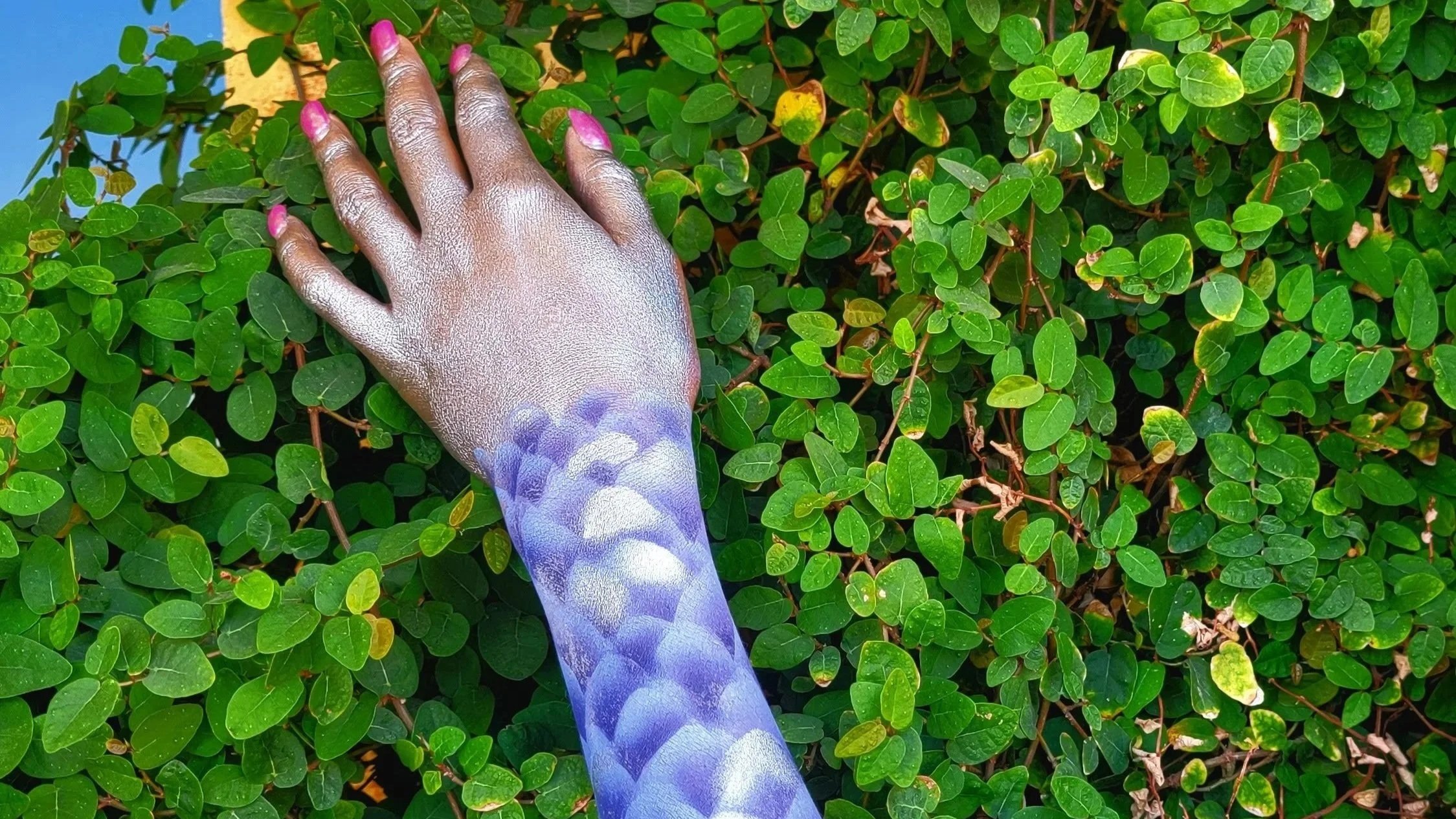 Professional body art and creative makeup services by Moore Beauty MT, featuring an arm transformation with intricate metallic silver and blue dragon scales against a background of lush green leaves.