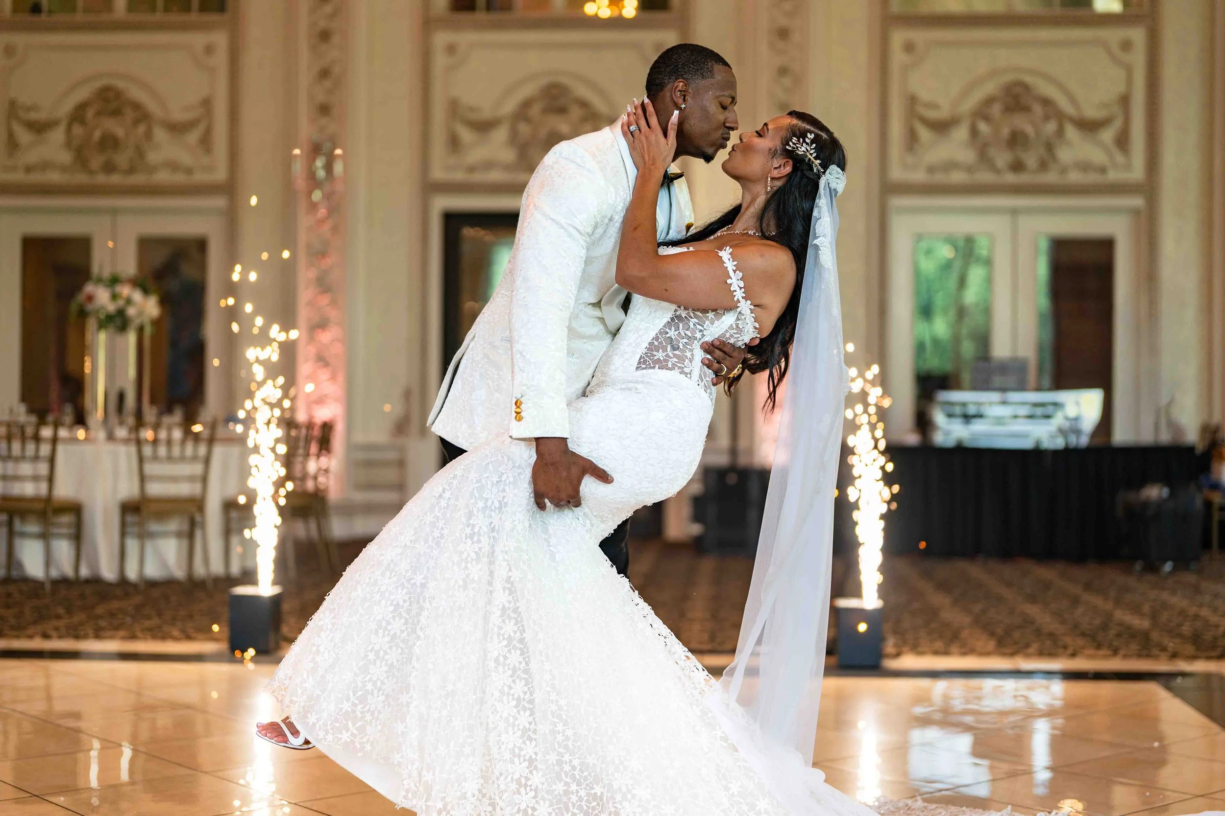 A bride and groom sharing a dance, with the groom lifting the bride in a ballroom decorated with chandeliers and sparklers.