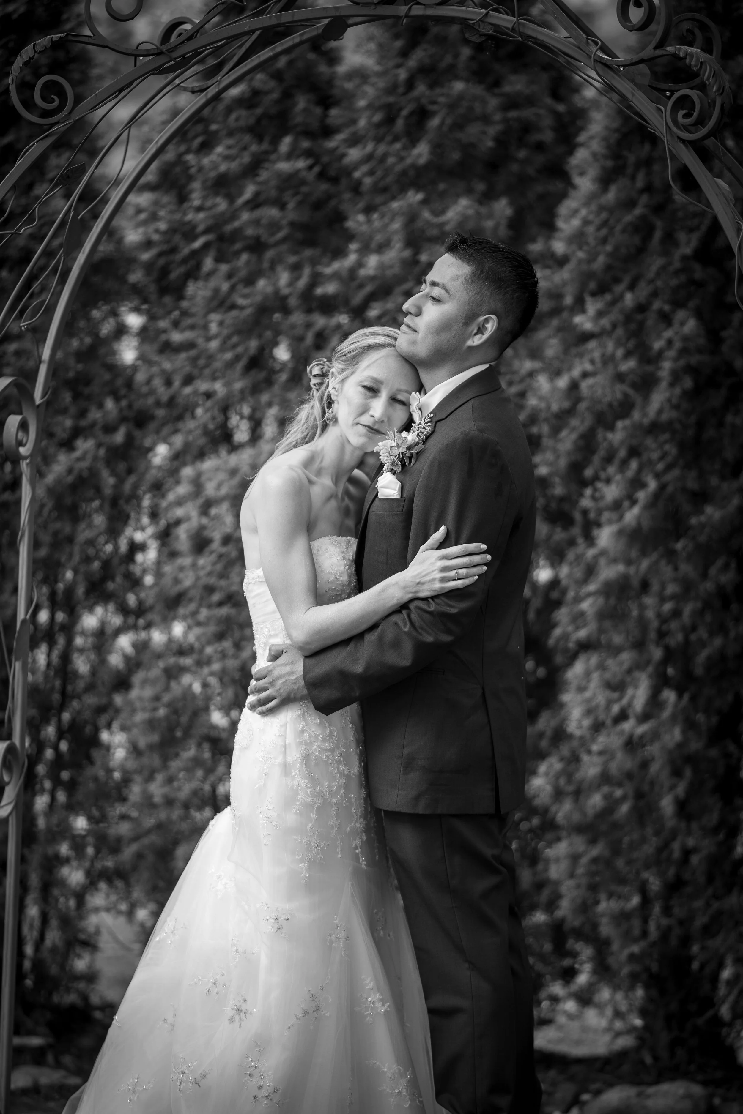 A black and white photograph of a bride and groom embracing under an ornate metal archway outdoors, with trees and foliage in the background.