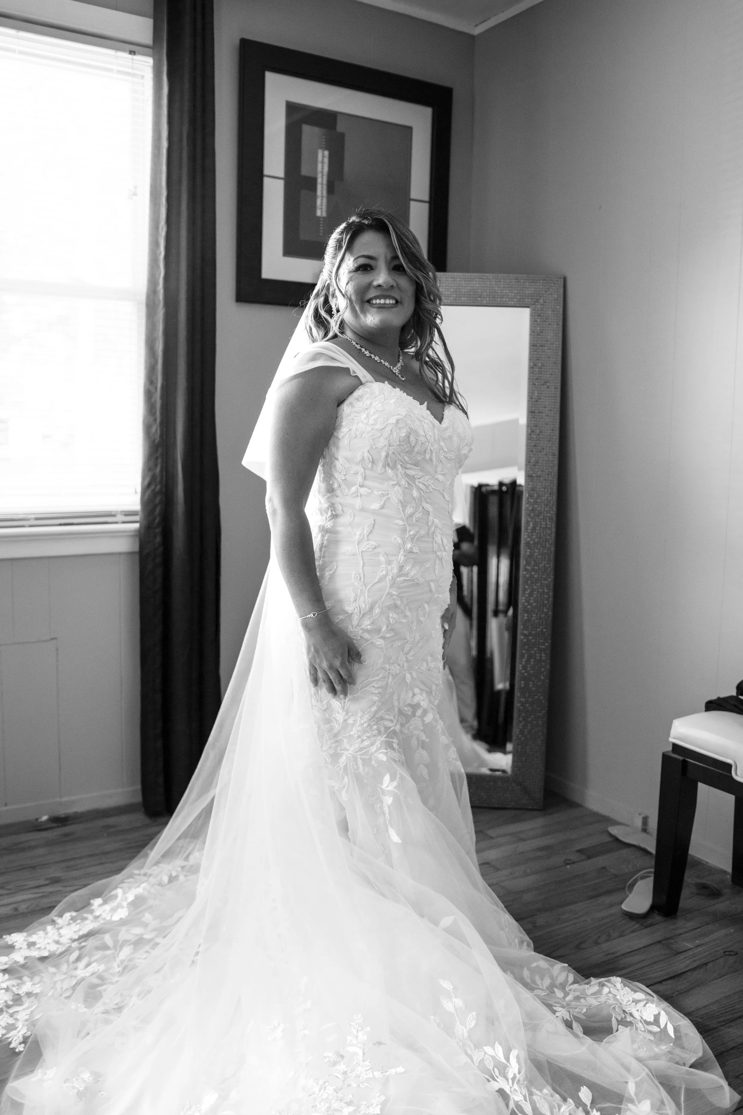 A woman in a wedding dress standing in a room with a mirror, smiling, with a window to her left and framed artwork on the wall behind her.