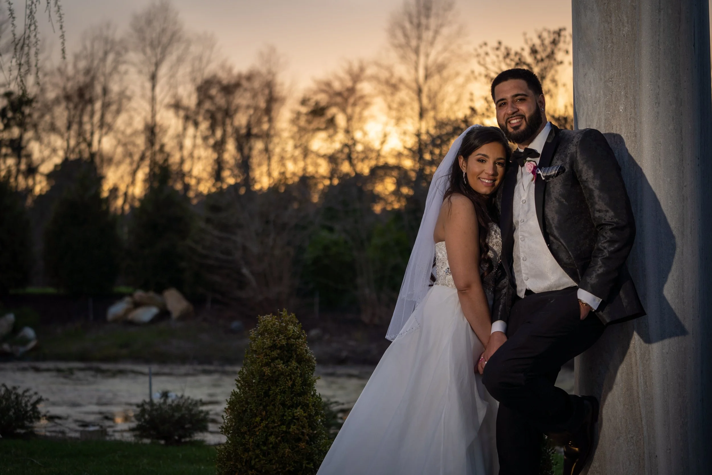 A newlywed couple is posing outdoors during sunset, standing close to a wall, smiling at the camera, with trees and a river in the background.
