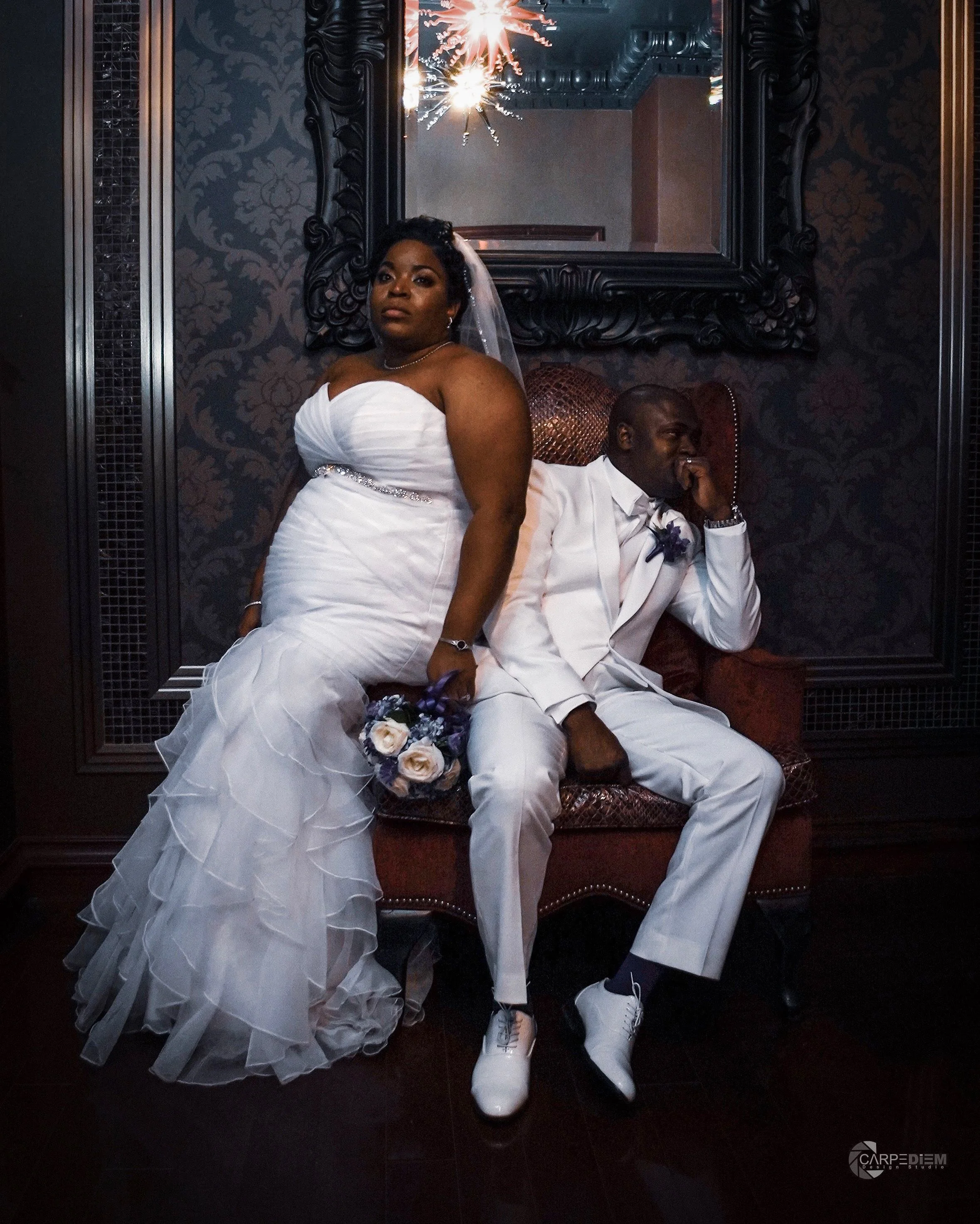 A bride in a white wedding gown holding a bouquet of purple and white roses, standing next to a man in a white suit sitting on a decorative chair. The setting is elegant with dark patterned wallpaper, a large ornate mirror, and sparklers reflecting i