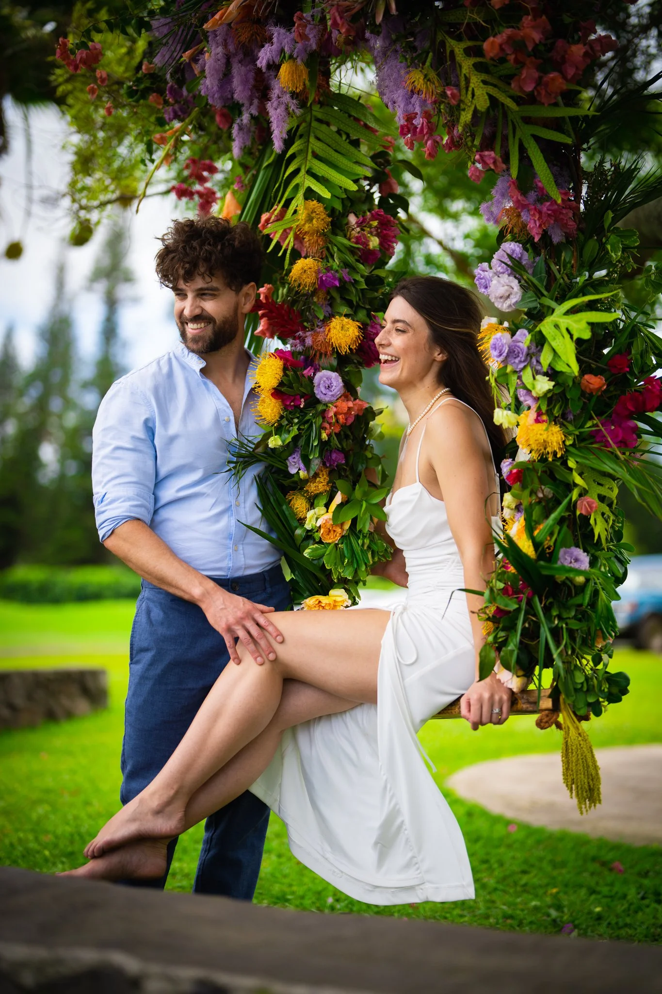 A couple on a decorated swing in a park, smiling and looking happy, surrounded by greenery and colorful flowers.