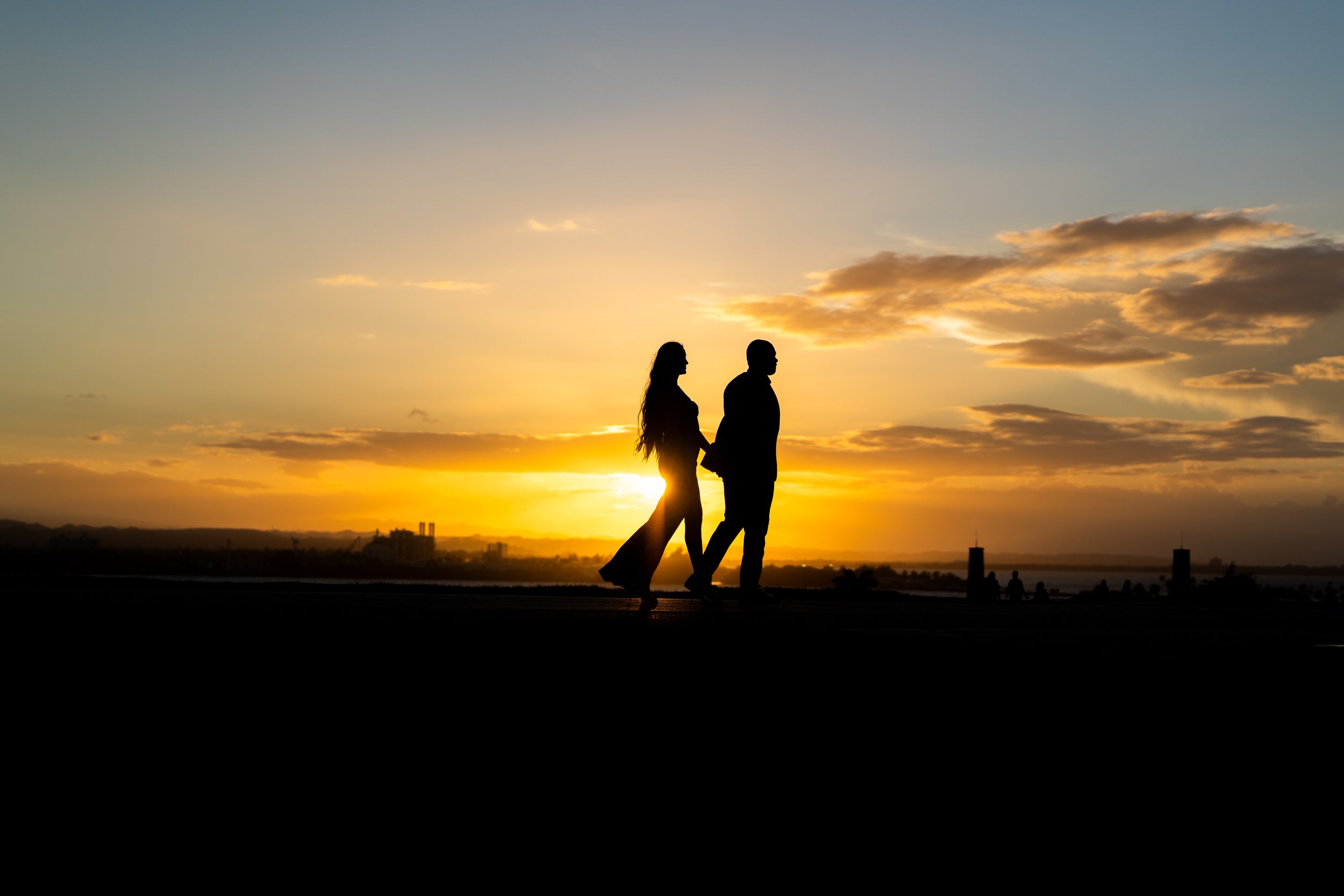 Silhouettes of a couple holding hands at sunset with a colorful sky and city skyline in the background.