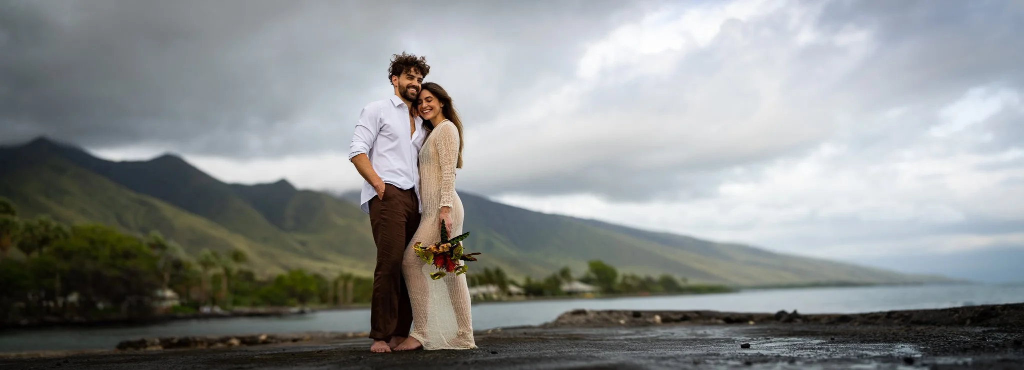 A couple standing close together on a beach with mountains in the background, the woman holding a bouquet of flowers, both smiling.