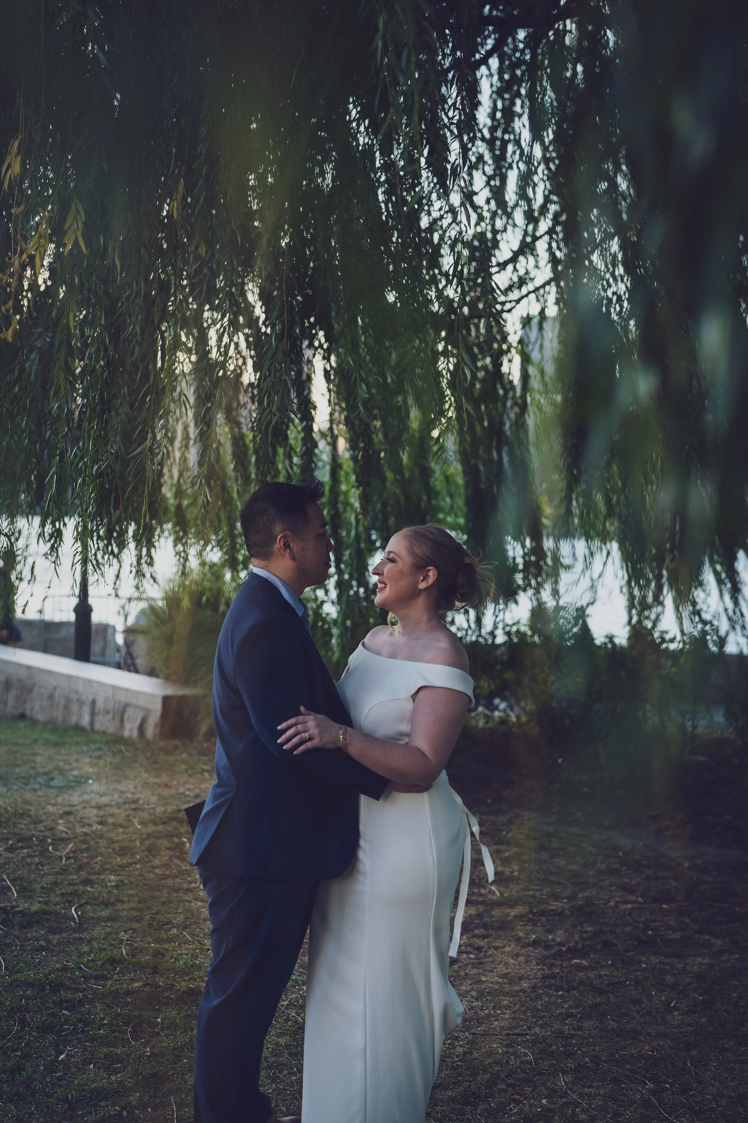 A wedding couple standing outdoors under a leafy tree, gazing into each other's eyes, with a body of water in the background.