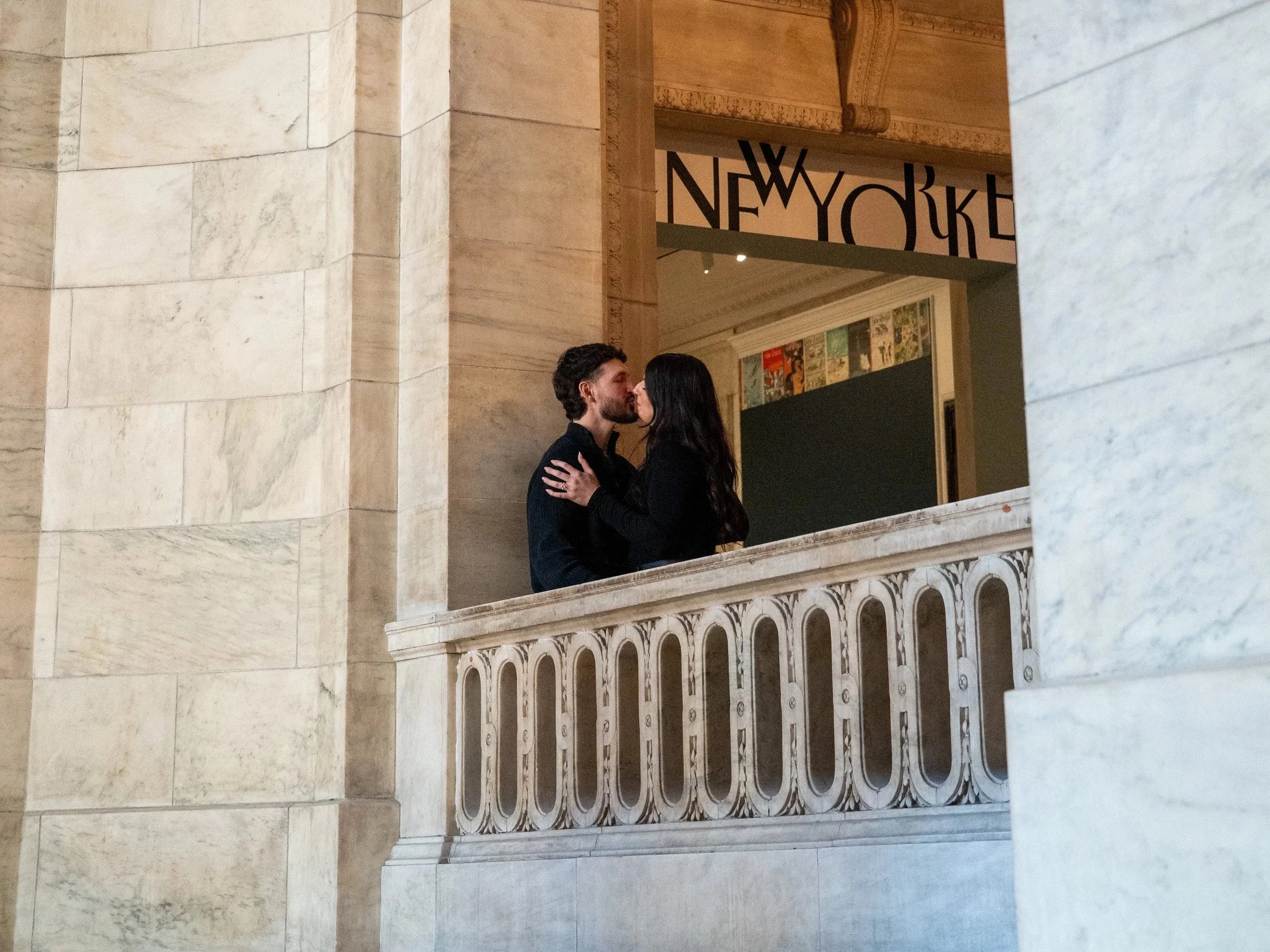 A couple kissing on a marble balcony inside a building with ornate architecture.