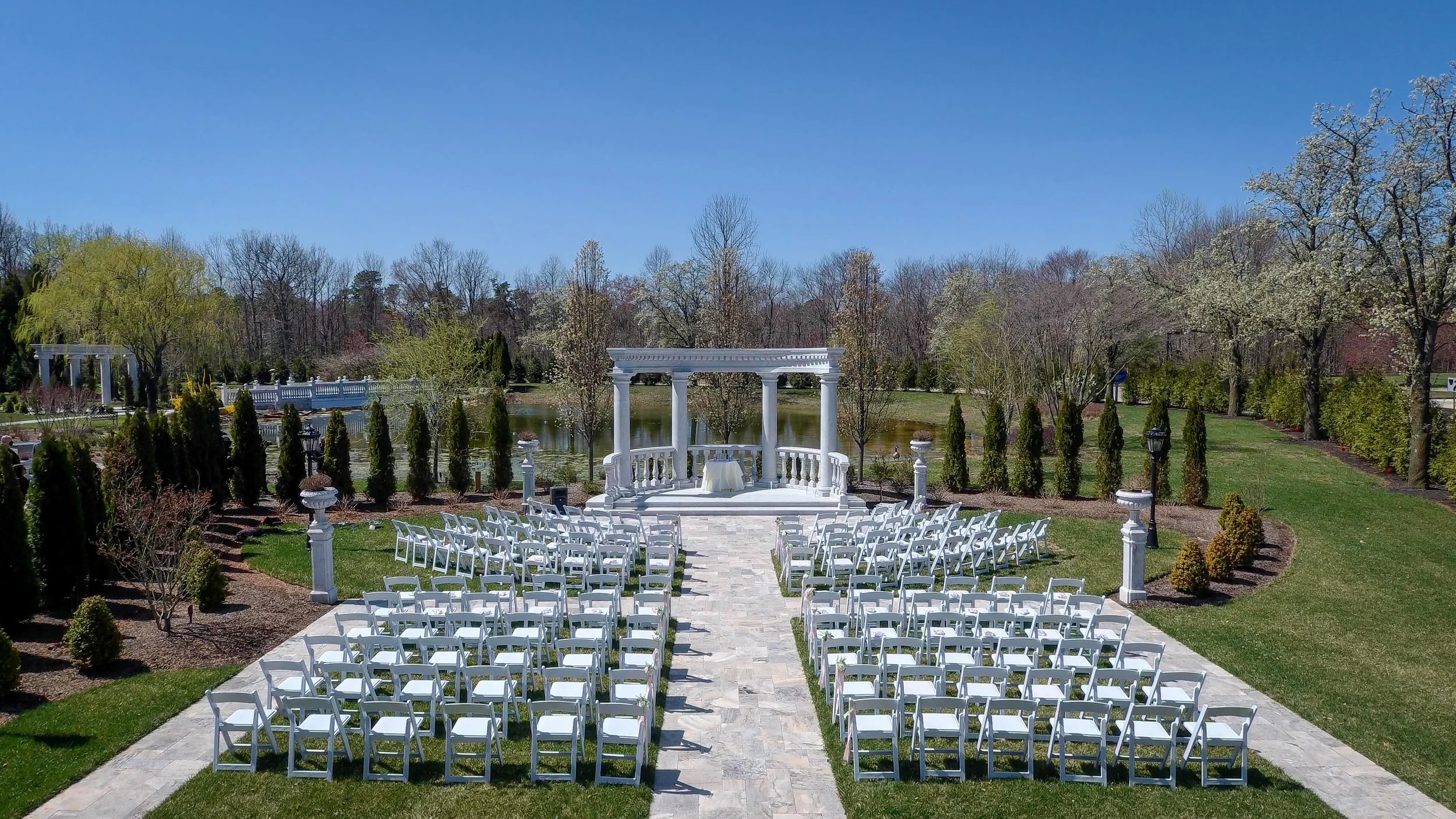 Outdoor wedding ceremony setup with white chairs arranged in rows on a stone path, facing a white classical pavilion near a pond, surrounded by trees and manicured bushes under a clear blue sky.