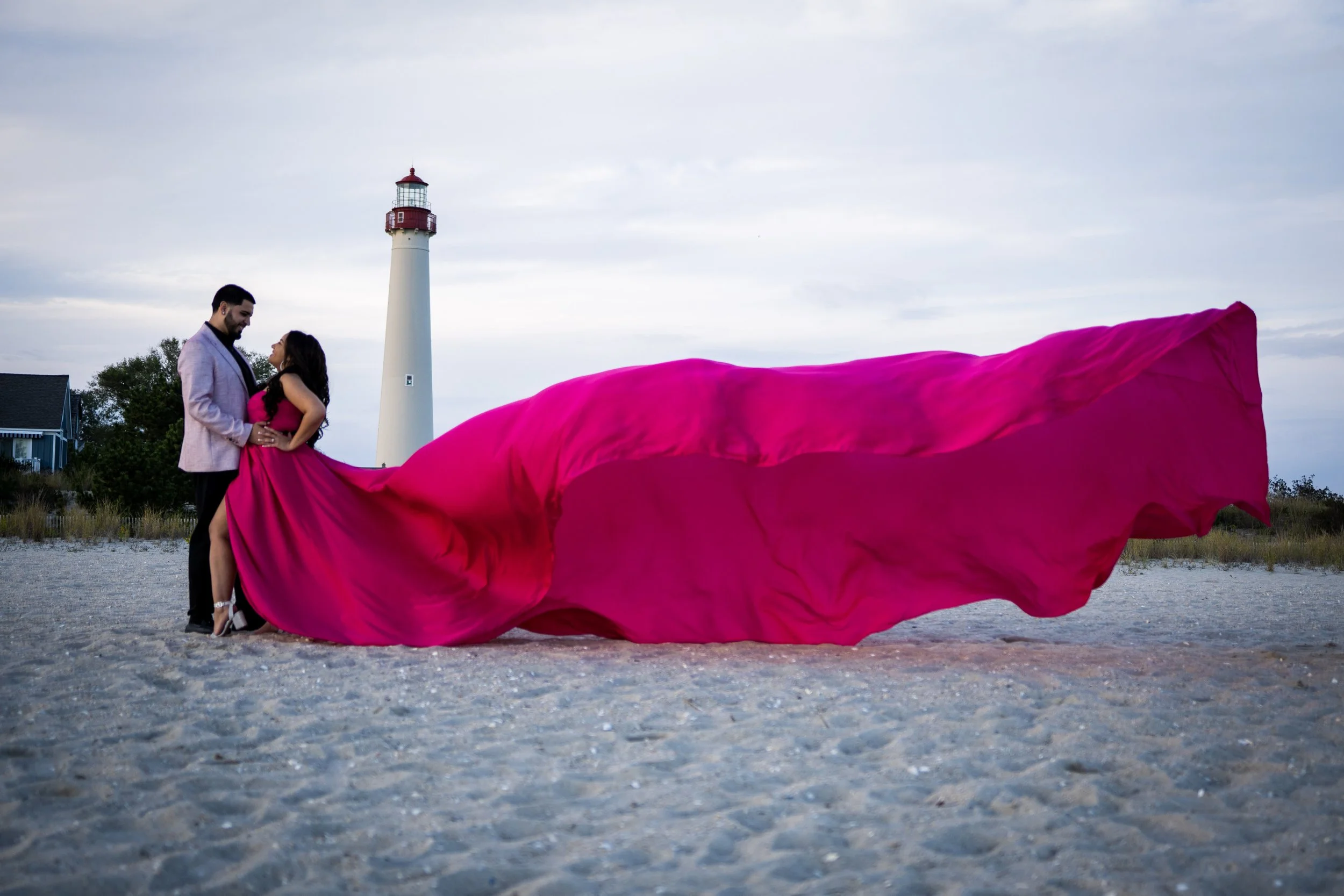 A couple dressed in formal attire standing on a sandy beach with a lighthouse in the background. The woman is wearing a long, flowing bright pink gown that extends onto the sand.