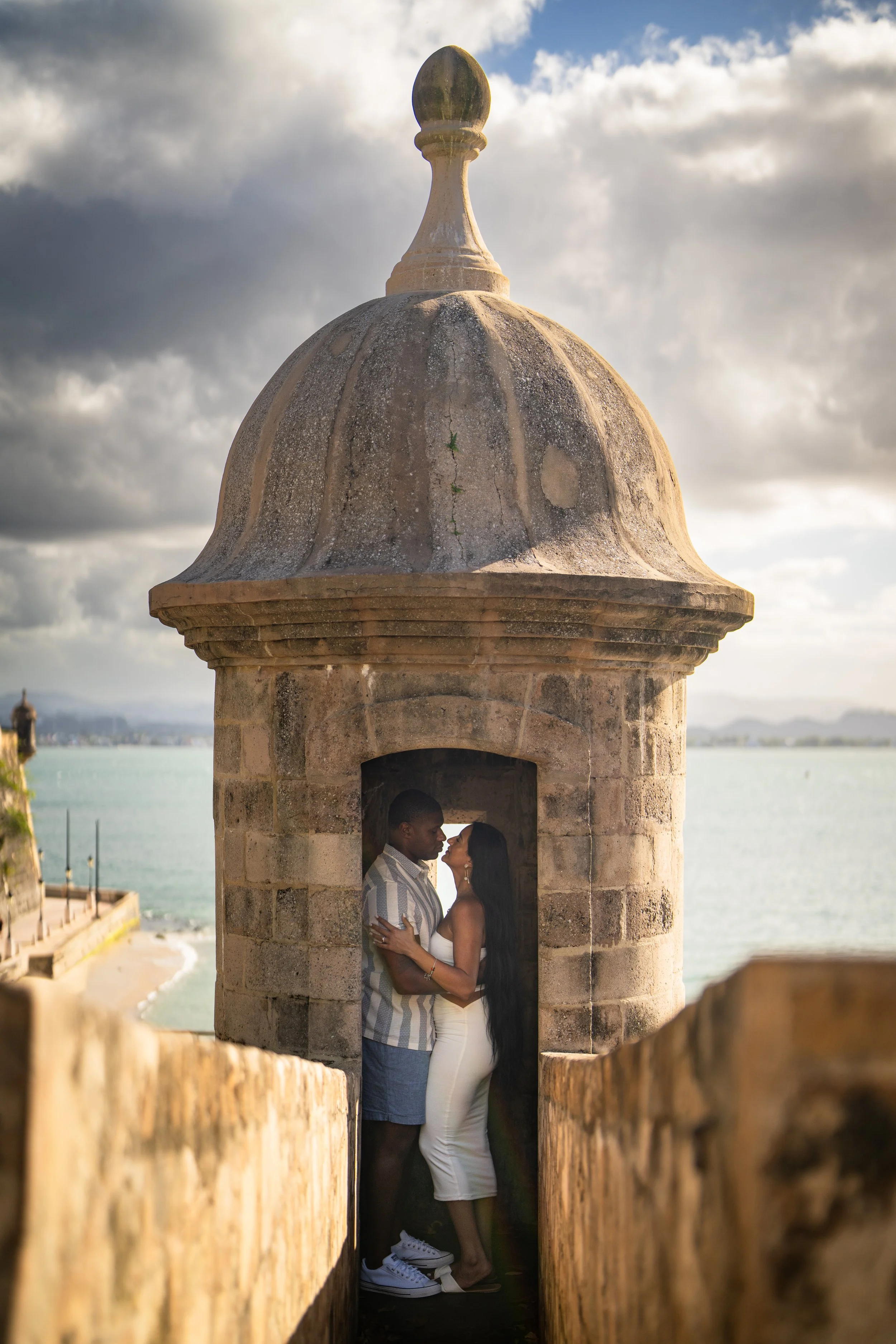 A couple standing close inside a small stone turret with a domed roof, overlooking the ocean, with cloudy sky in the background.