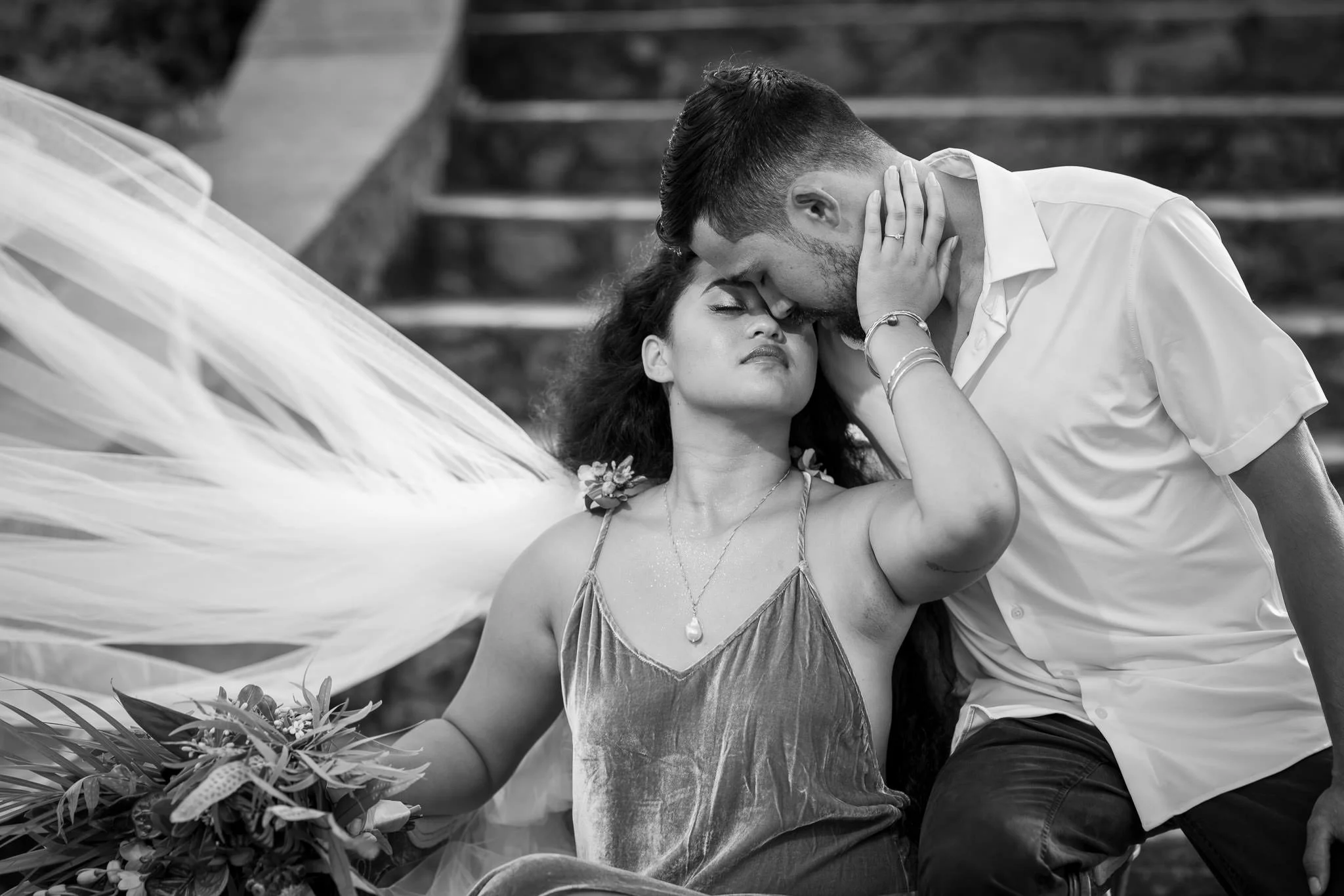 A black and white photograph of a man and woman in an intimate pose, with the man gently kissing the woman's forehead as she closes her eyes. The woman has flowers in her hair and is holding a bouquet, while the man is leaning in, touching her face with his hand.