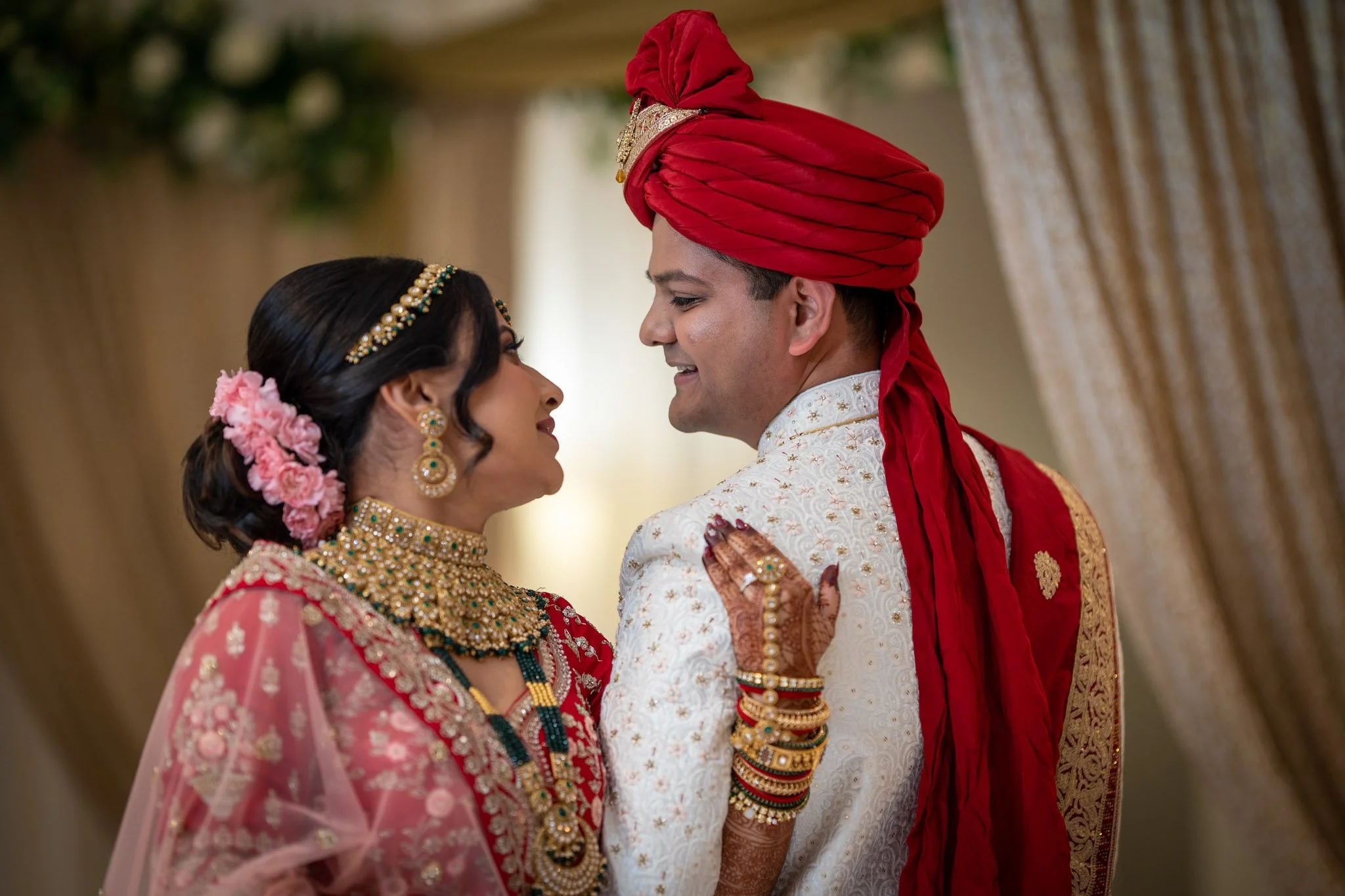 A bride and groom in traditional Indian wedding attire, facing each other lovingly. The bride is wearing a pink sari, elaborate jewelry, and has flowers in her hair. The groom is dressed in a white sherwani with a red turban.