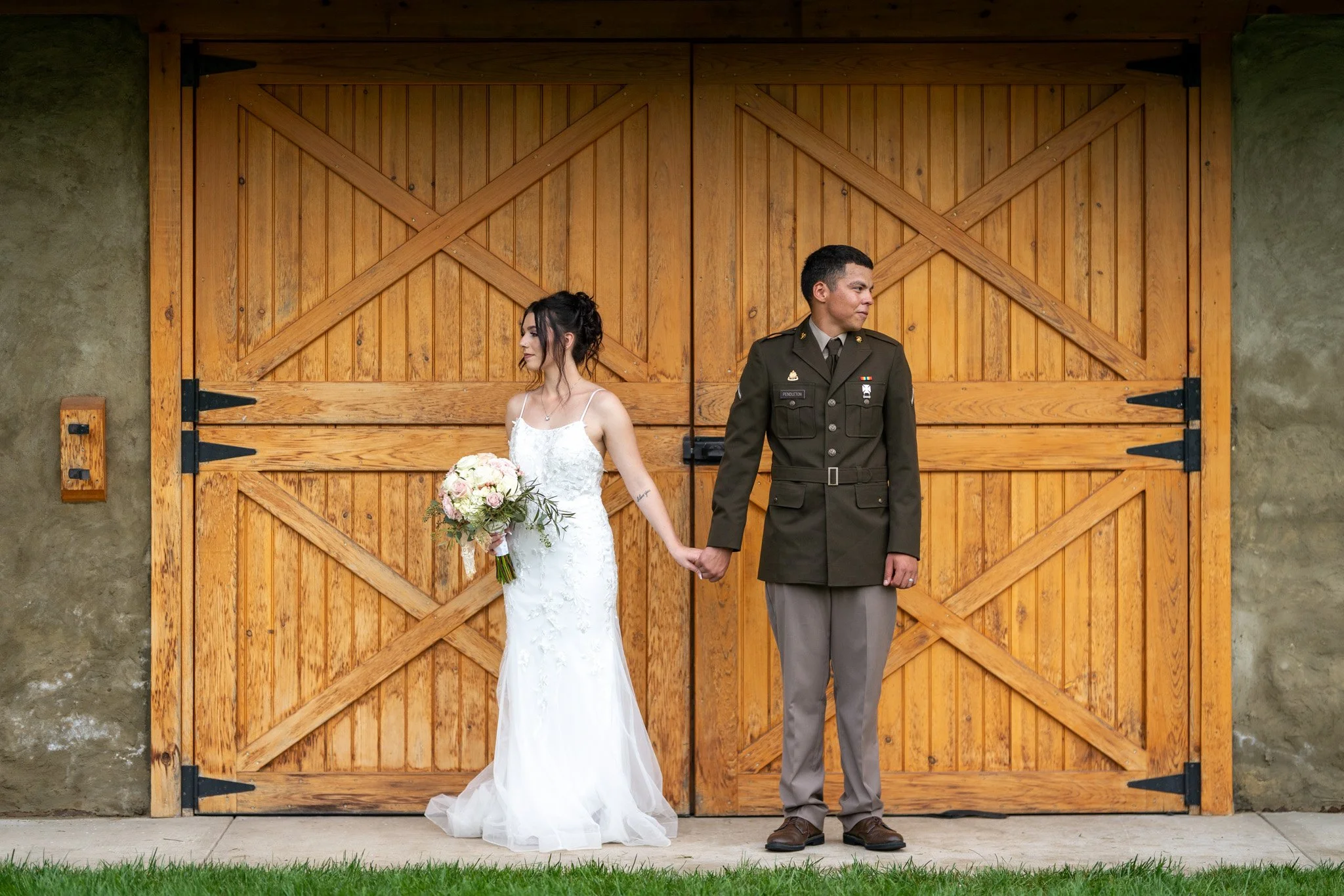 A bride in a white wedding dress holding a bouquet of flowers and a groom in a military uniform hold hands and stand apart in front of a wooden barn door, looking away from each other.
