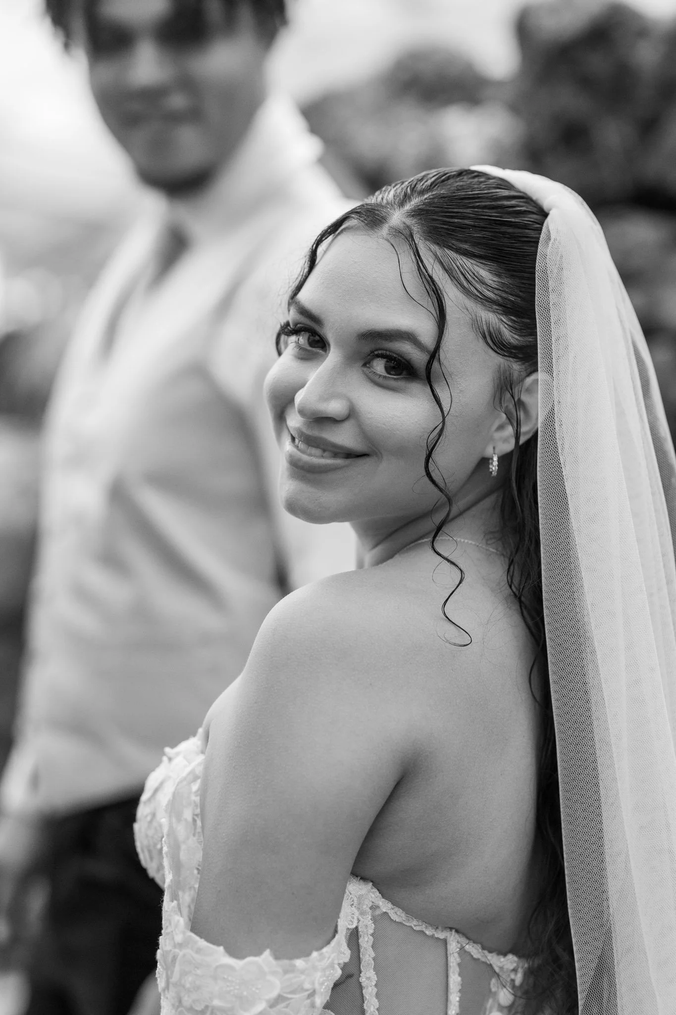 A smiling bride with curly hair and a veil, looking over her shoulder, with a groom blurred in the background.