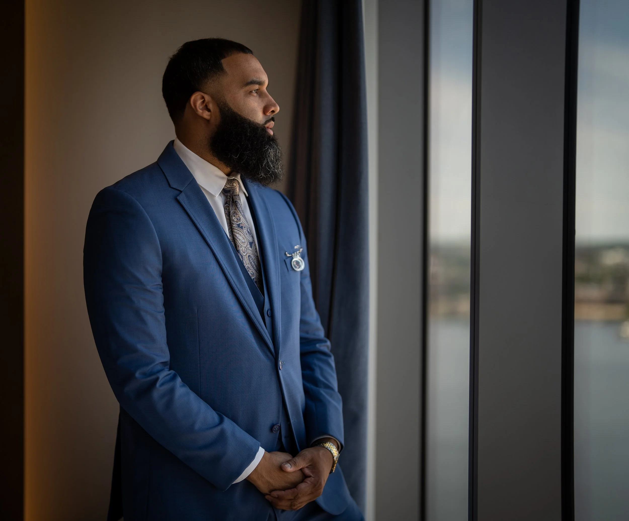Man in a blue suit and tie standing by a window looking outside.