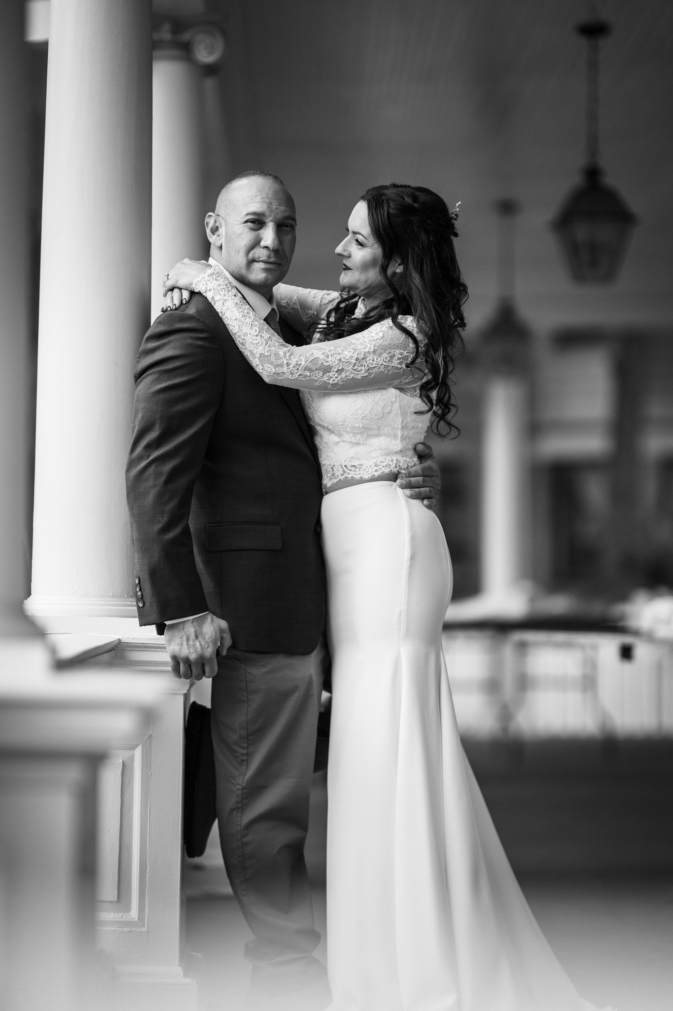 Black and white photo of a man in a suit and a woman in a wedding dress embracing indoors near large white columns and hanging lanterns.