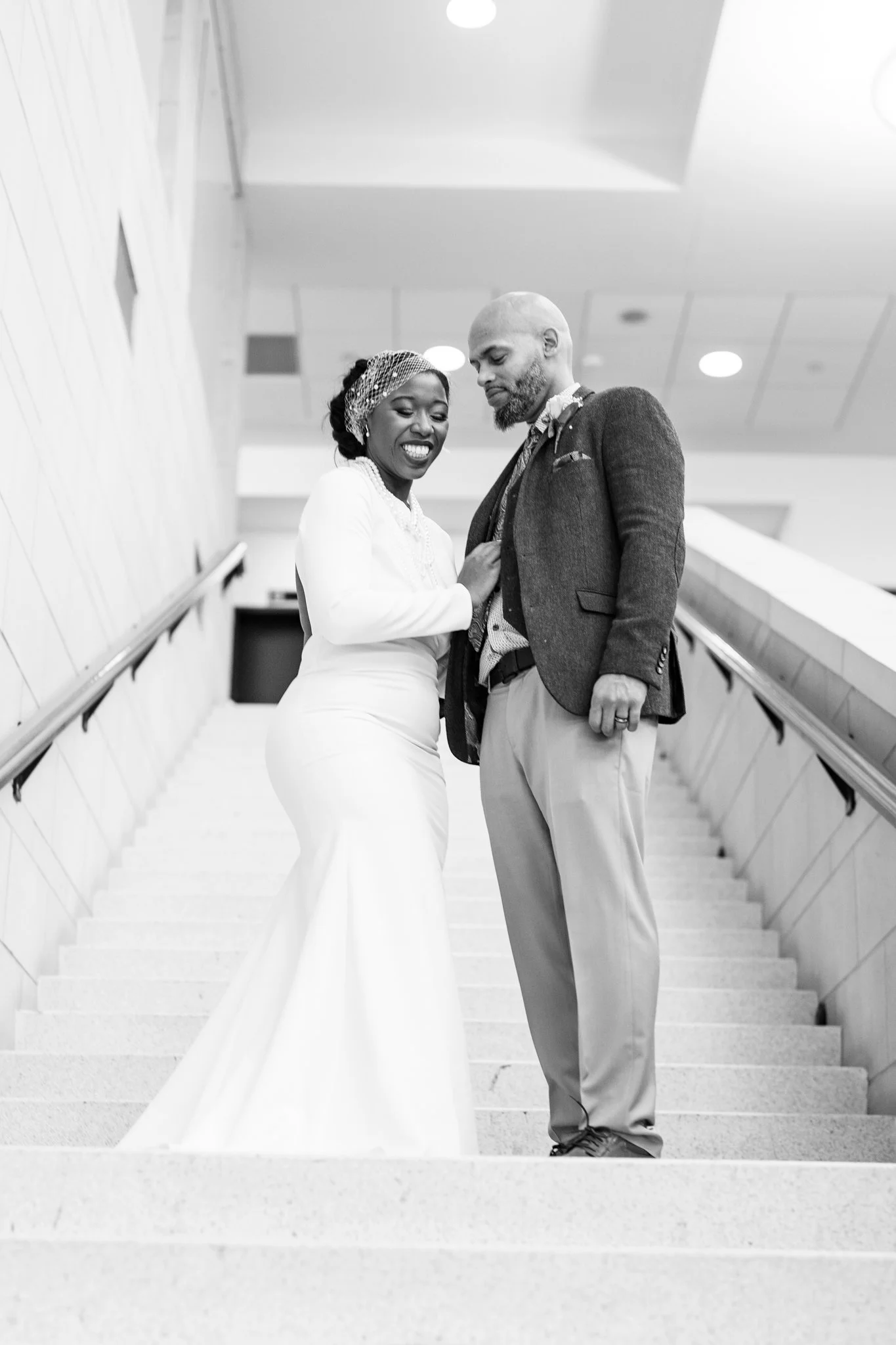 Black and white photo of a happy woman in a long white wedding gown and headband, and a man in a suit, standing close together on staircase in an indoor setting.