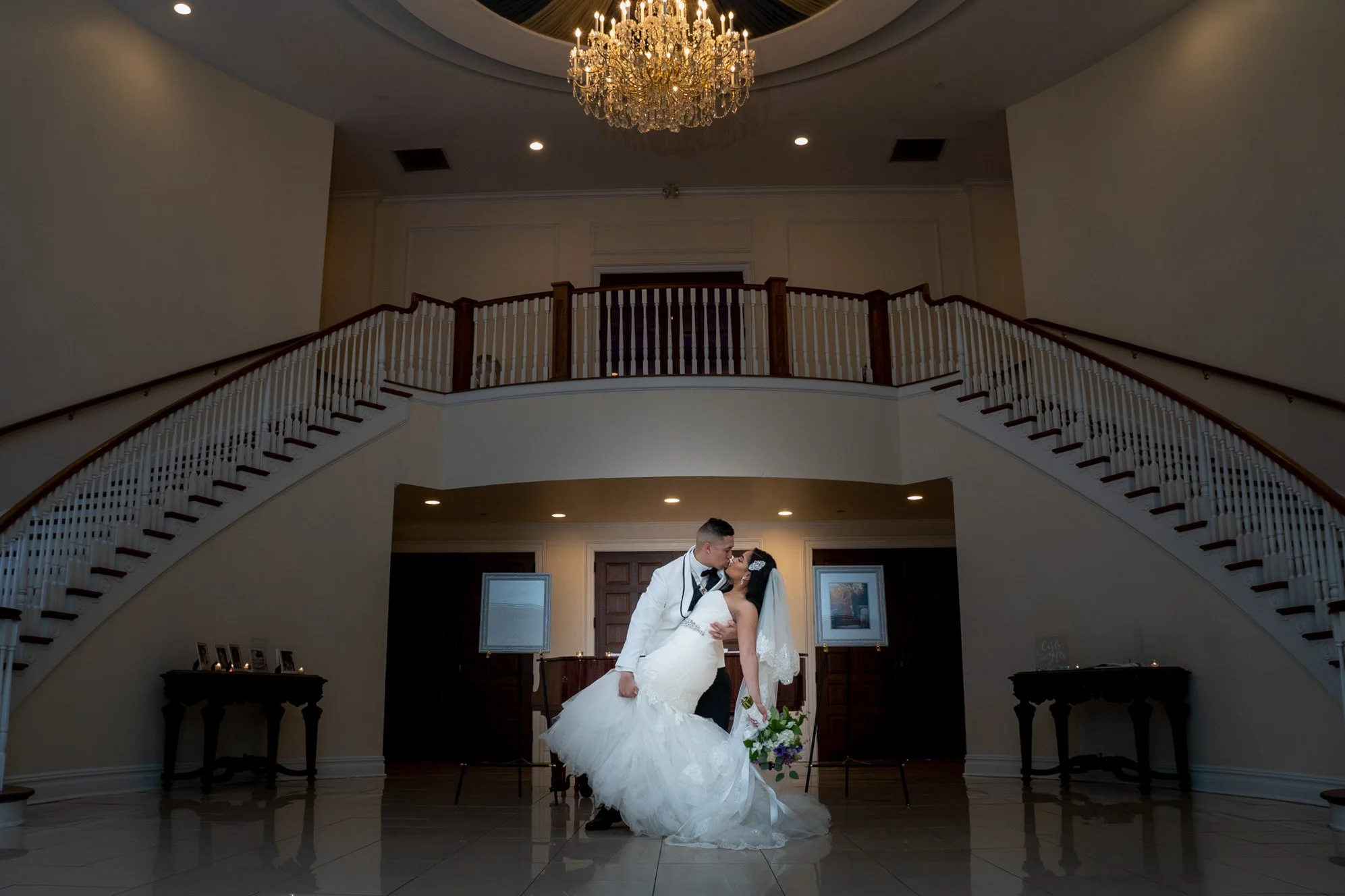 A bride and groom stand close, kissing in an elegant, spacious room with a grand staircase and chandelier overhead. The bride wears a white wedding dress and veil, holding a bouquet, while the groom is dressed in a tuxedo.