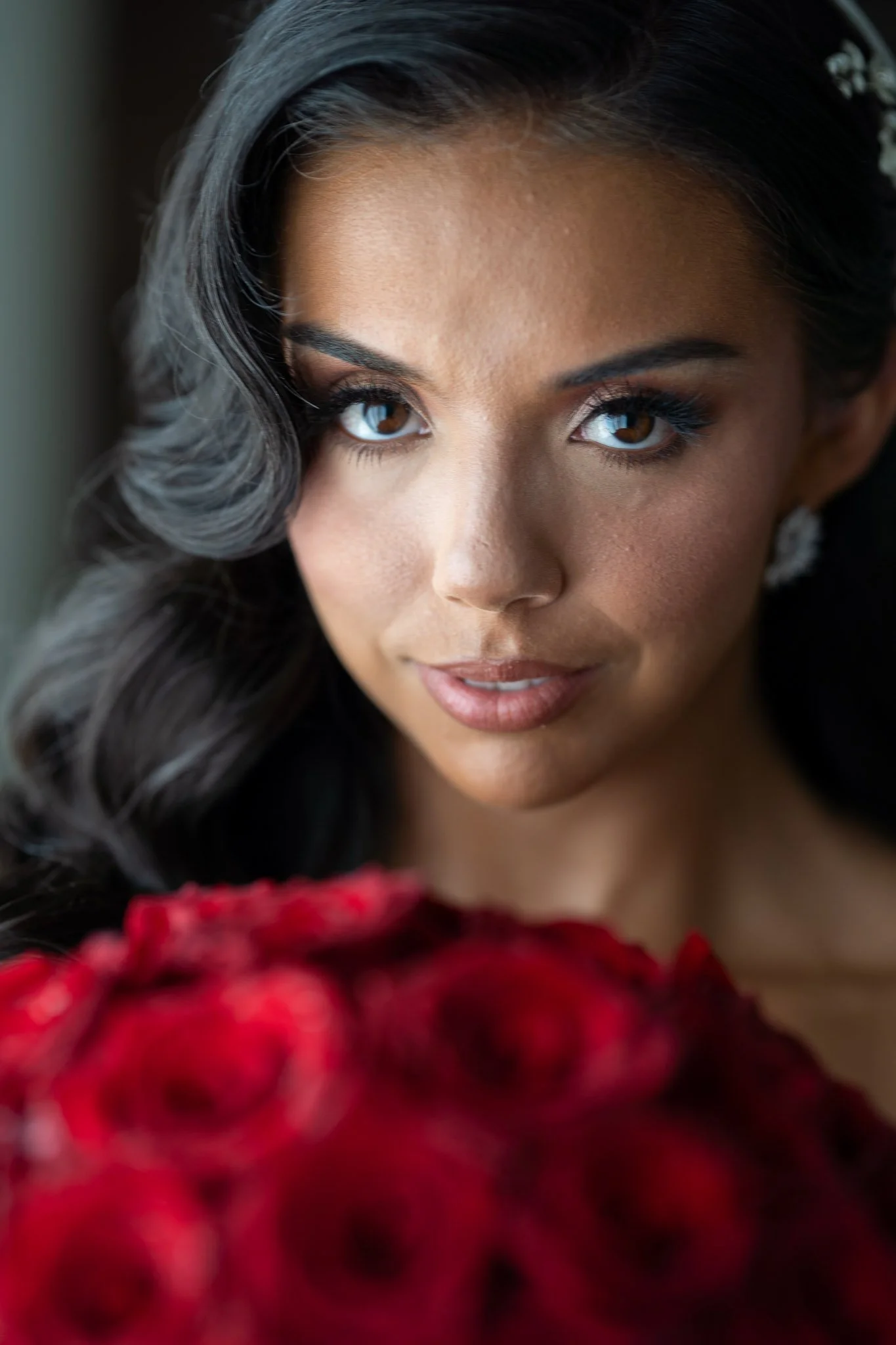 Close-up of a woman with dark hair and brown eyes, looking directly at the camera, holding a bouquet of red roses.