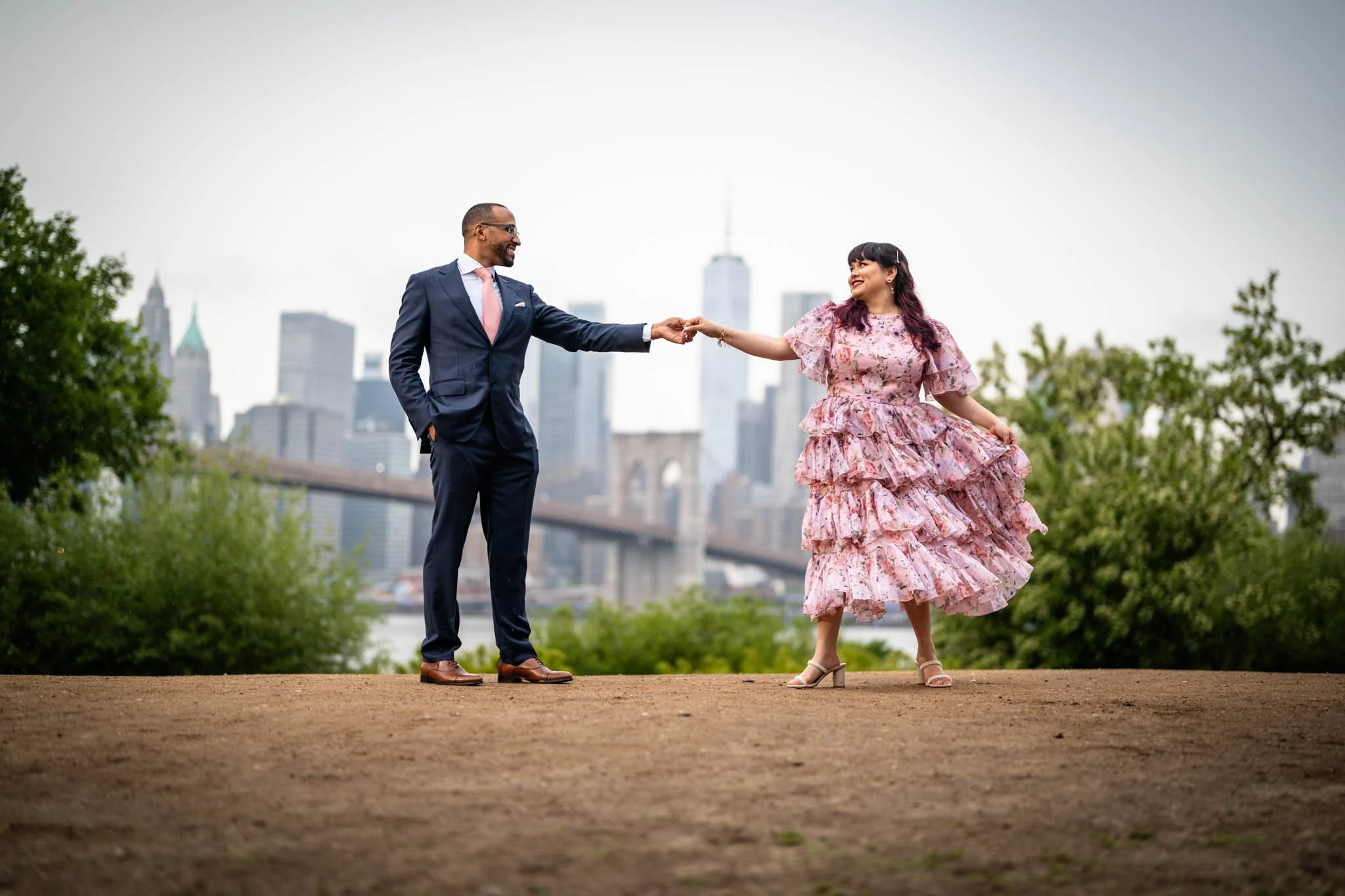 A man and woman holding hands and smiling outdoors, with New York City skyline and Brooklyn Bridge in the background.