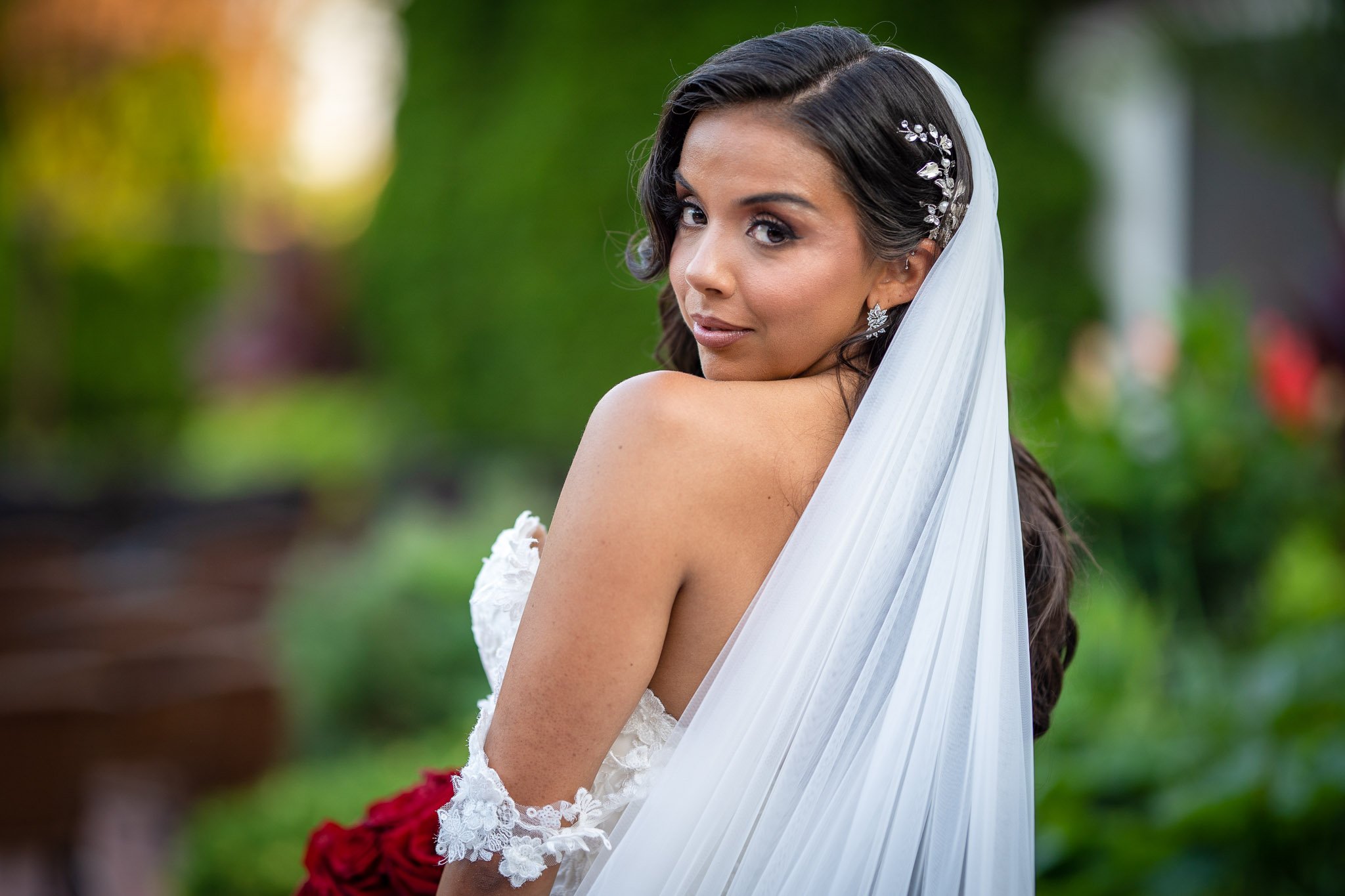 A bride with dark hair, styled in loose waves with a decorative hairpiece, wearing a strapless white wedding gown with lace details, holding a bouquet of red roses, outdoors with a blurred green background.