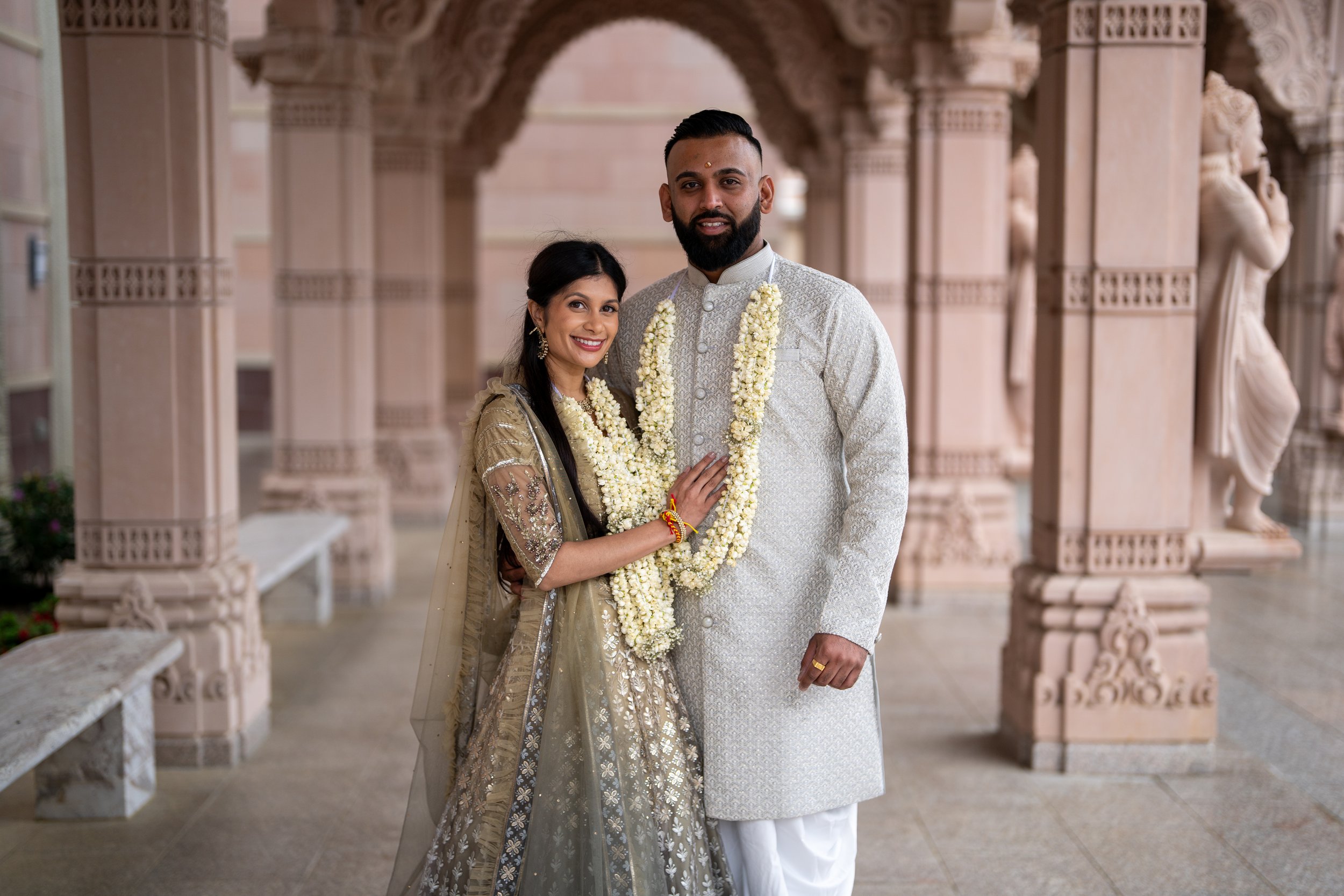 A South Asian couple dressed in traditional wedding attire standing in a courtyard with carved pink stone columns and statues in the background. The groom is wearing a white sherwani with a garland, and the bride is in an embellished gold and silver 