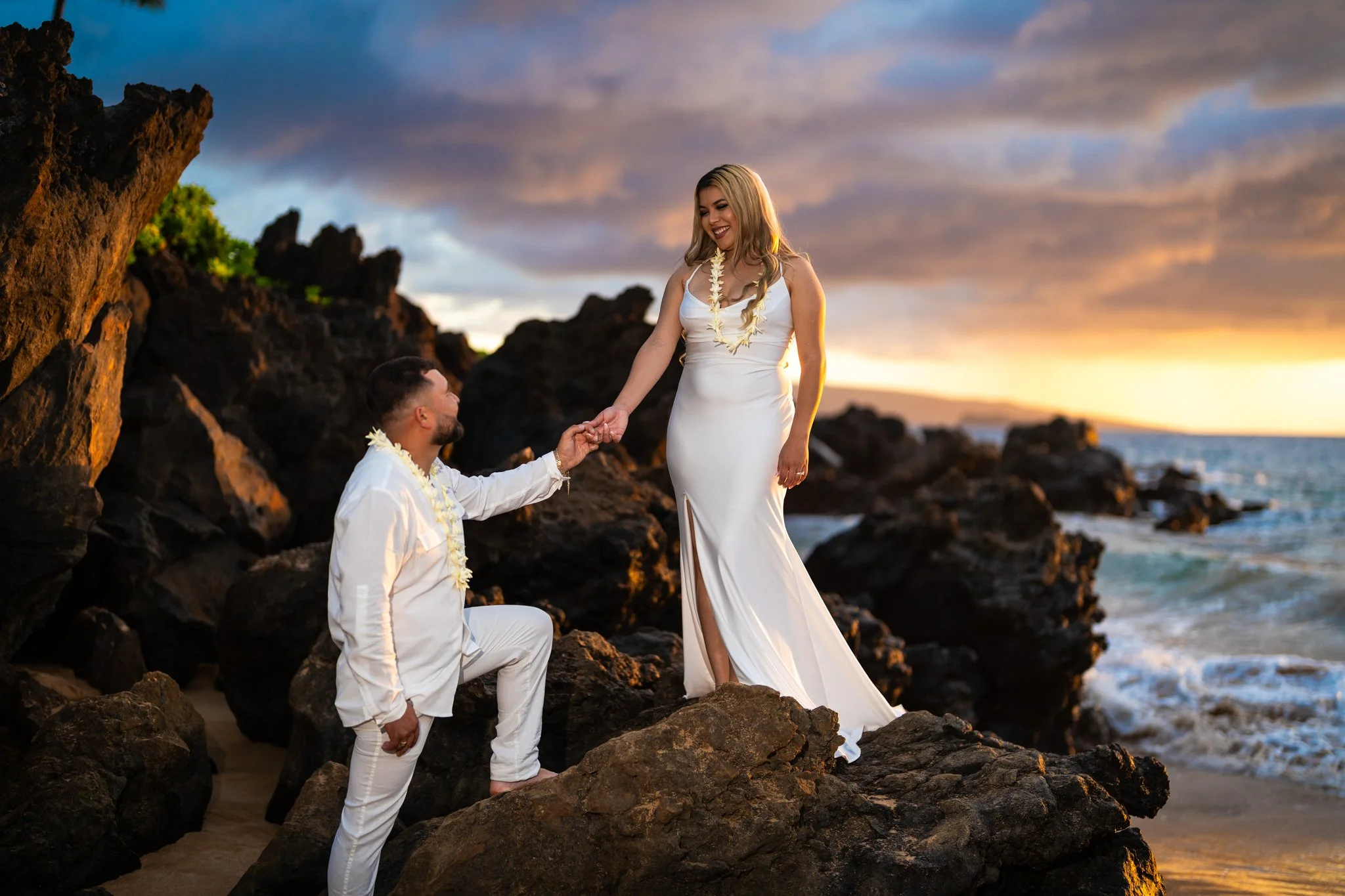 A couple in wedding attire on a rocky beach at sunset. The woman in a white gown is standing on a rock and smiling, holding hands with the man who is kneeling and reaching out to her. The sky is partly cloudy with a golden sunset.