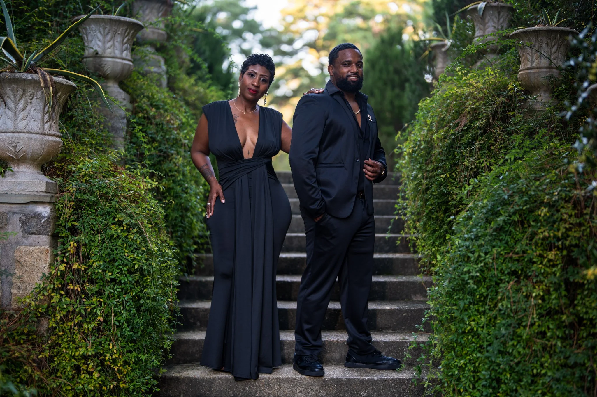 A man and woman dressed in formal black attire standing on outdoor stone steps, surrounded by lush green bushes and tall planter pots. The woman is wearing a long, deep V-neck dress, and the man is wearing a suit with his hand in his pocket.