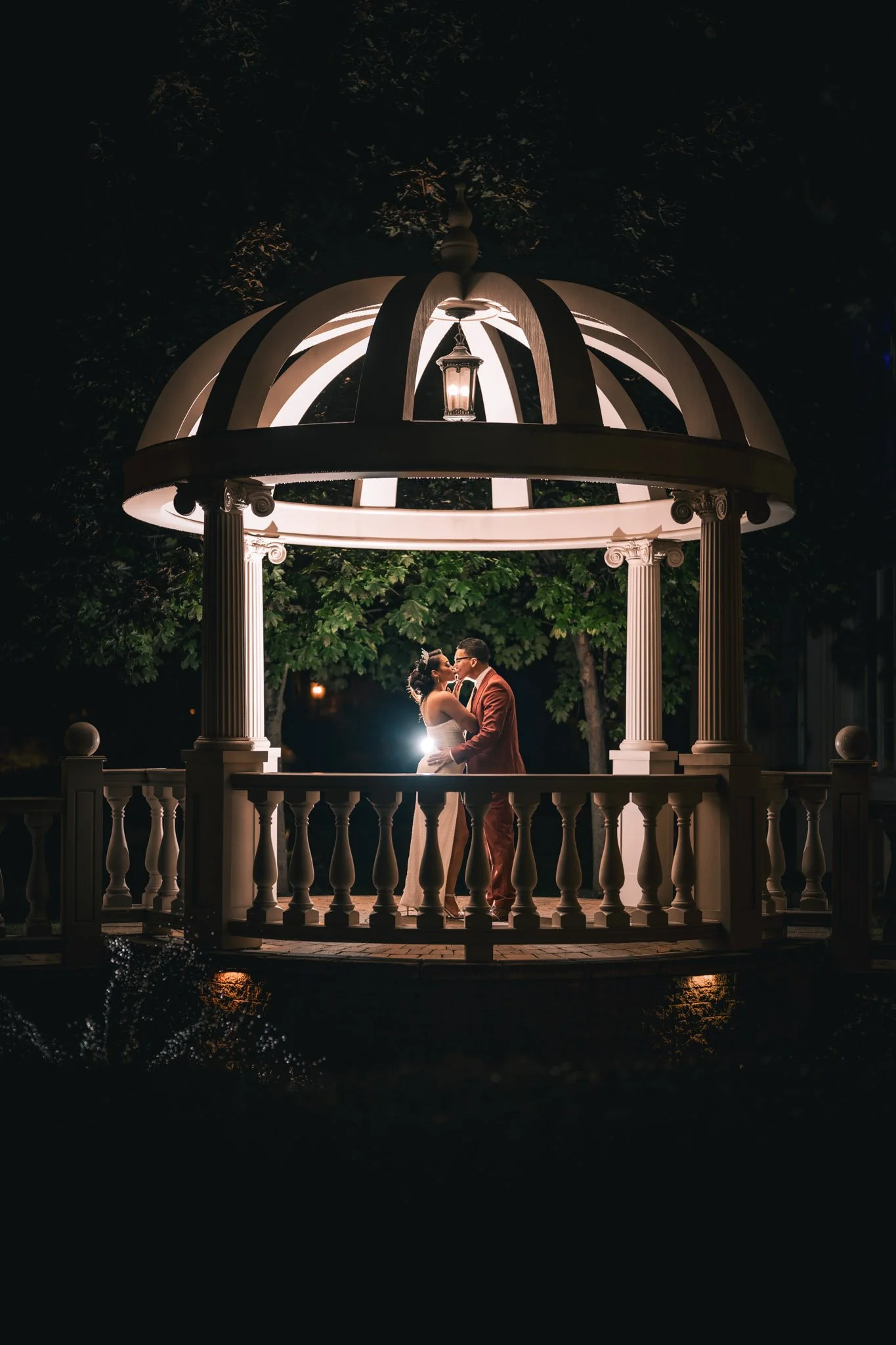 A couple in formal attire sharing a kiss under a gazebo at night, illuminated by a backlight creating a romantic scene with trees in the background.