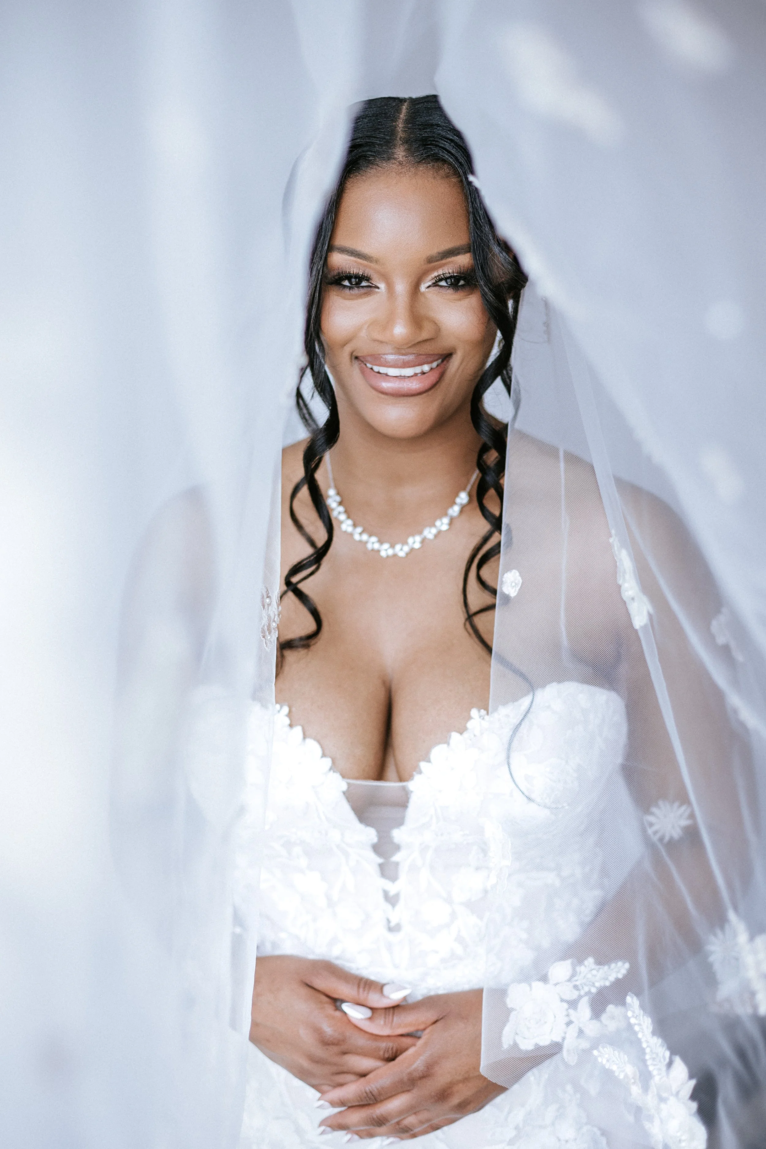 A woman in a bridal gown, smiling, with dark wavy hair and jewelry, partly covered by her wedding veil.