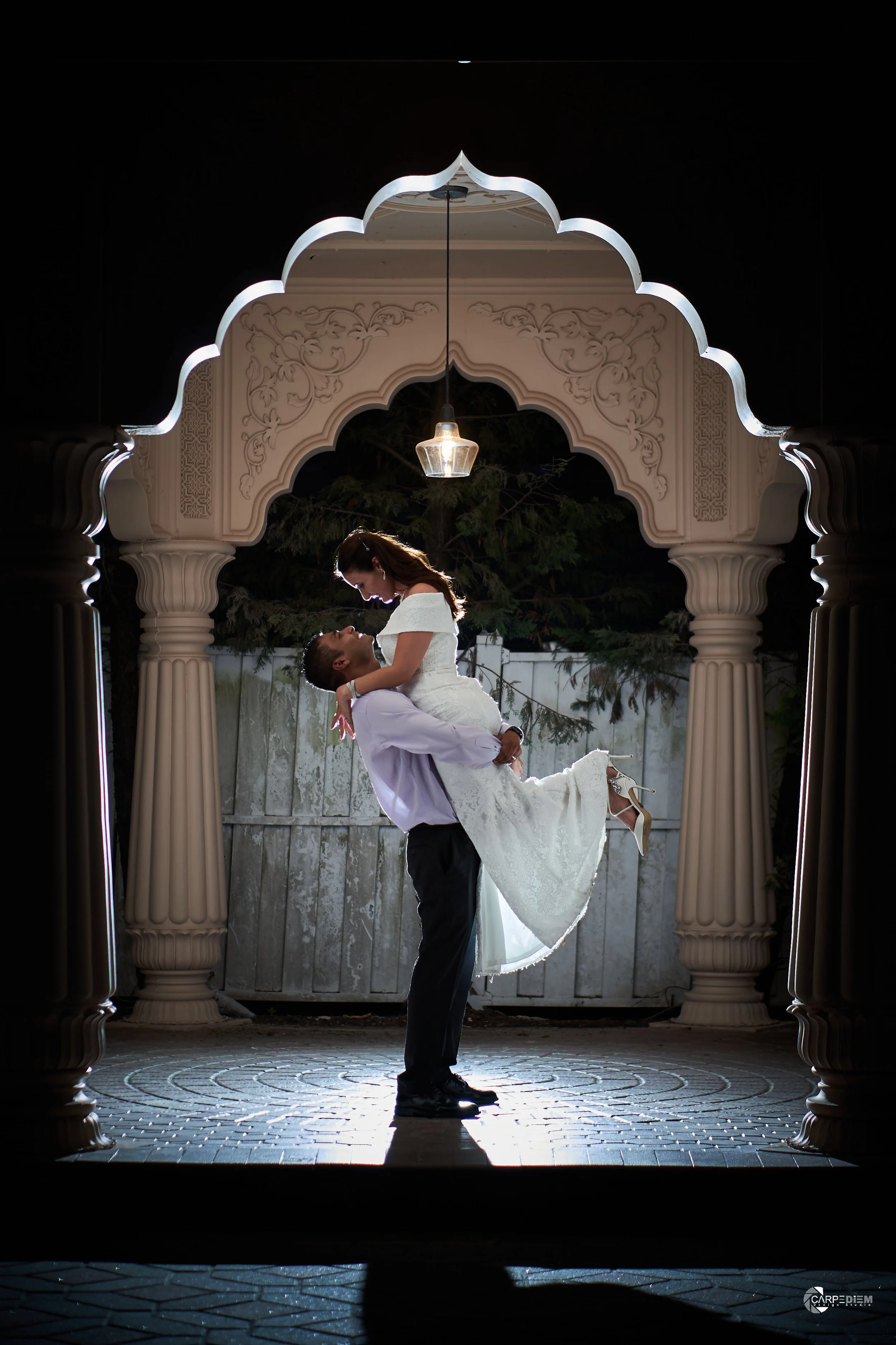 A groom lifting a bride under a decorative archway at night, with a hanging light above them and a wooden fence in the background.