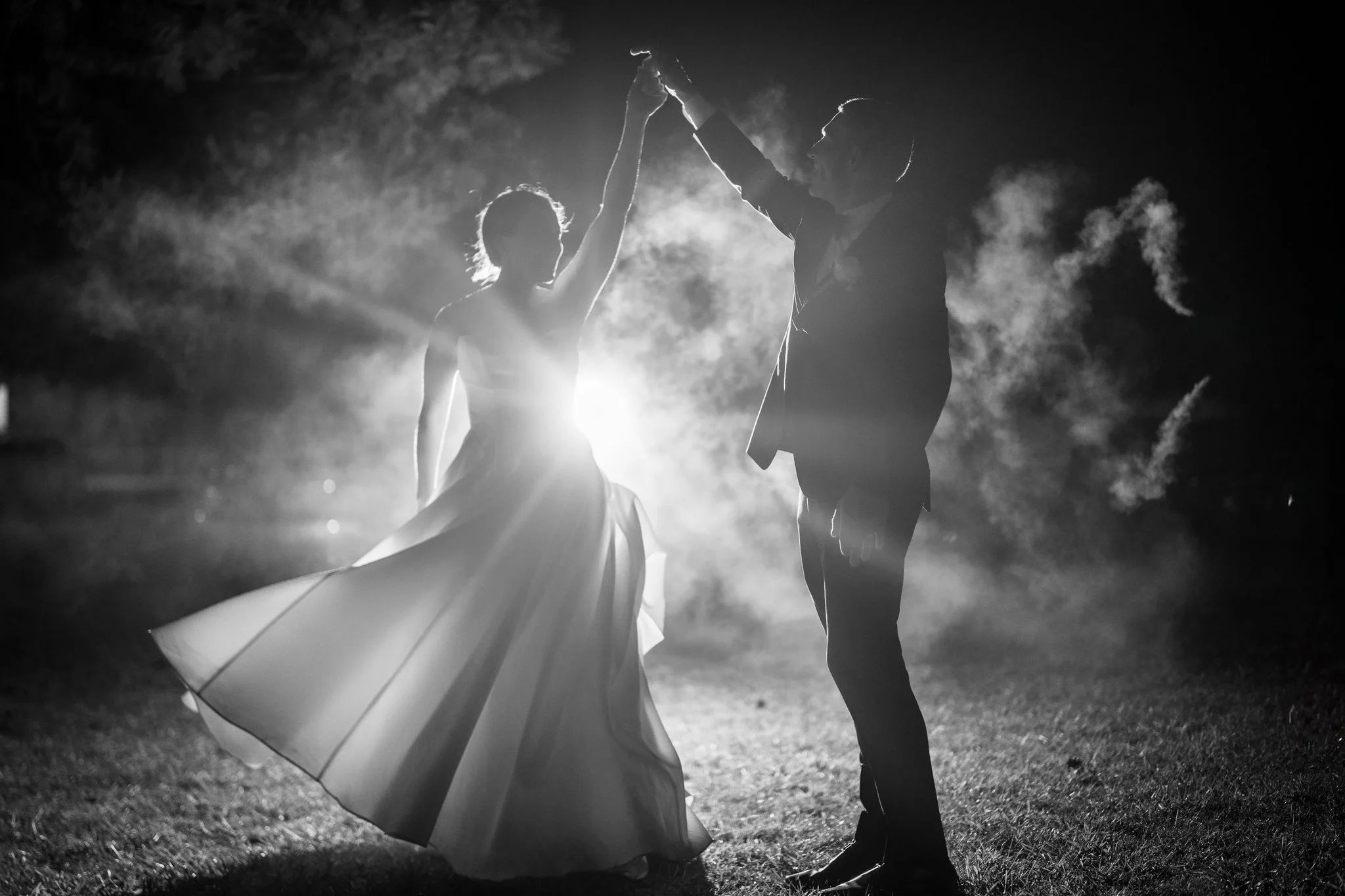 A couple dancing outdoors at night, backlit with a bright light behind them, creating a silhouette effect in black and white.