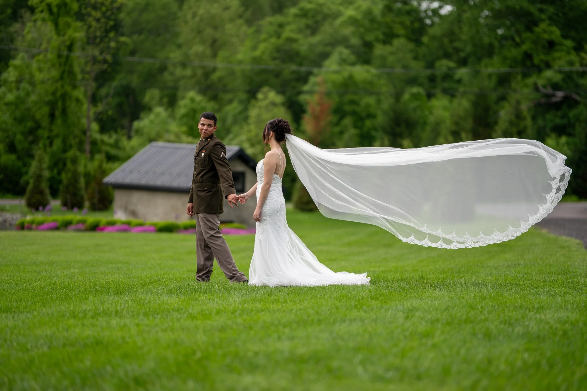A bride in a white wedding dress and a groom in a military uniform holding hands on a grassy field with a small building and green trees in the background.