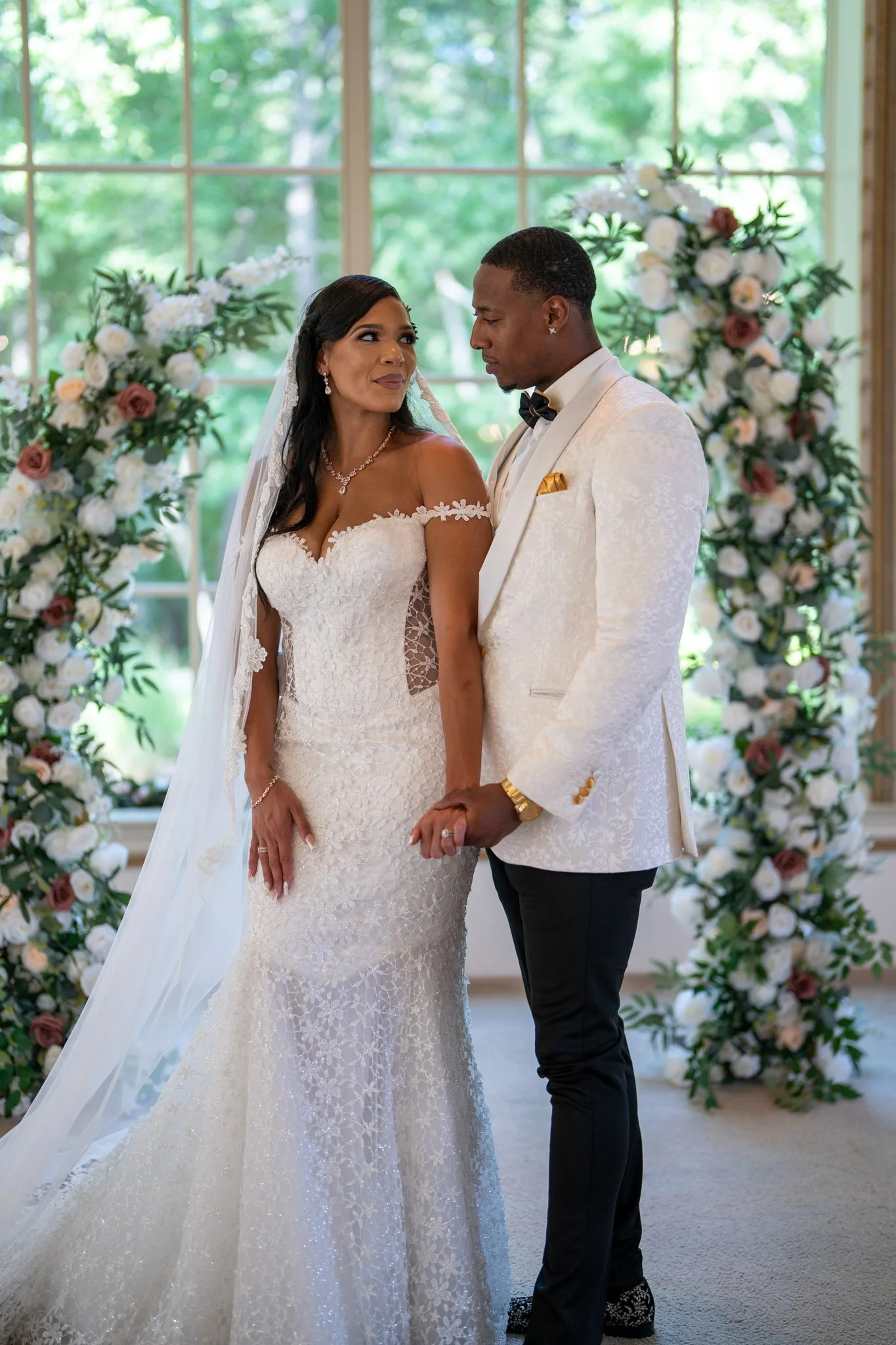 A bride and groom holding hands during their wedding ceremony, standing in front of floral arch with white and pink roses, inside a bright room with large windows showing green trees outside.