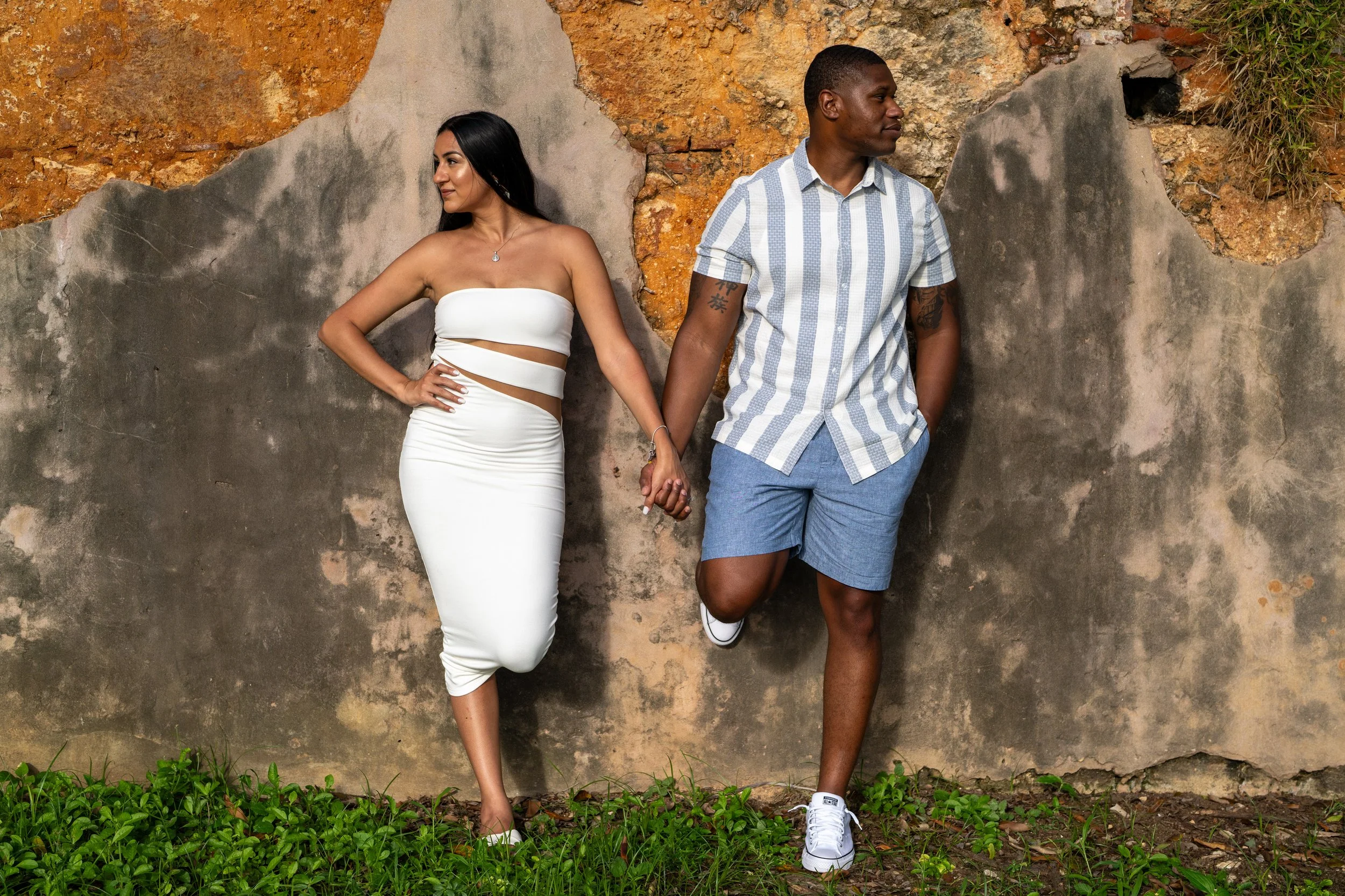 A couple holding hands, standing against a textured wall. The woman is wearing a white, strapless dress with cutouts, and the man is dressed in a short-sleeved, striped shirt and blue shorts.