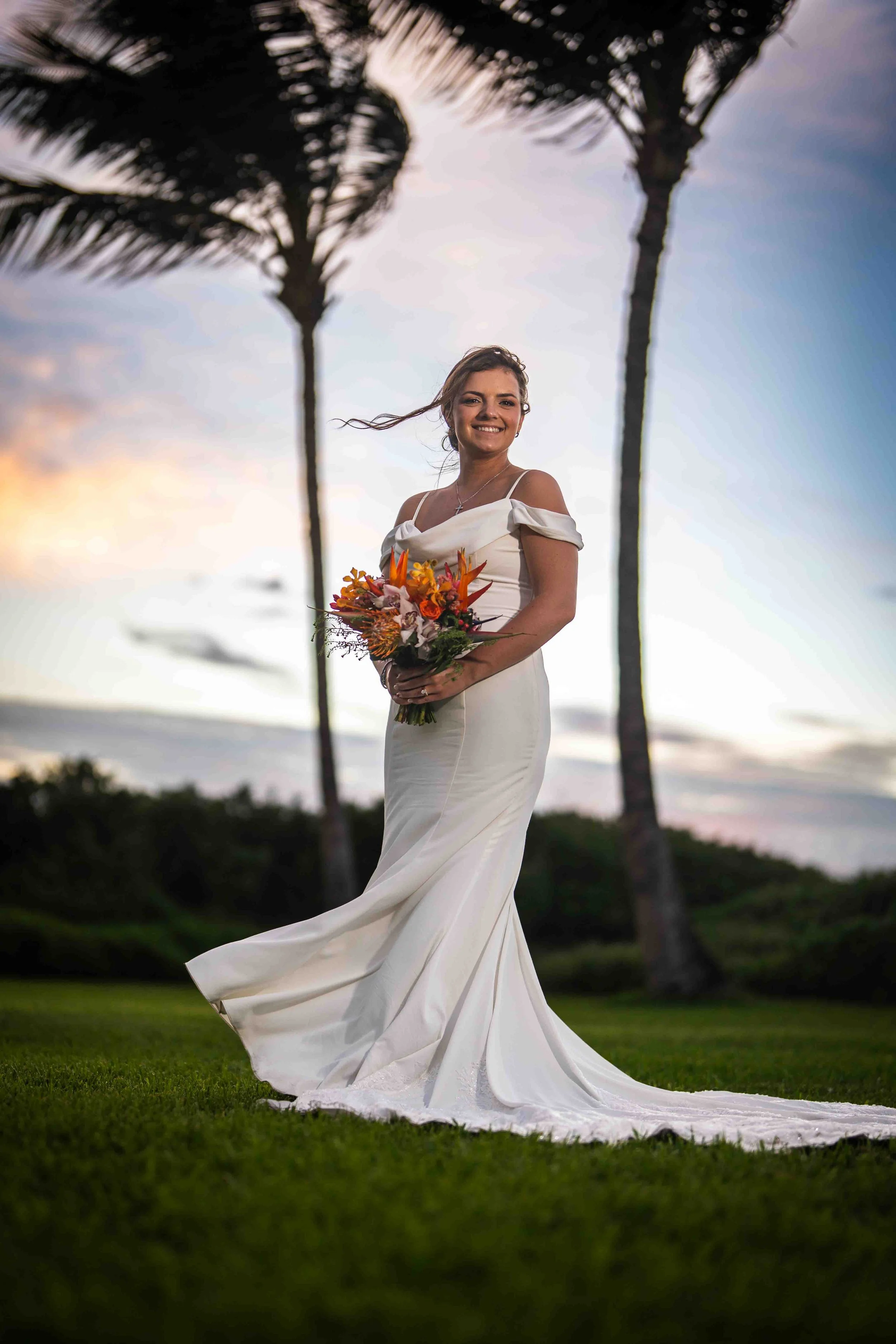 A bride in a white wedding dress holding a bouquet of orange, pink, and red flowers outdoors at sunset, with palm trees in the background.