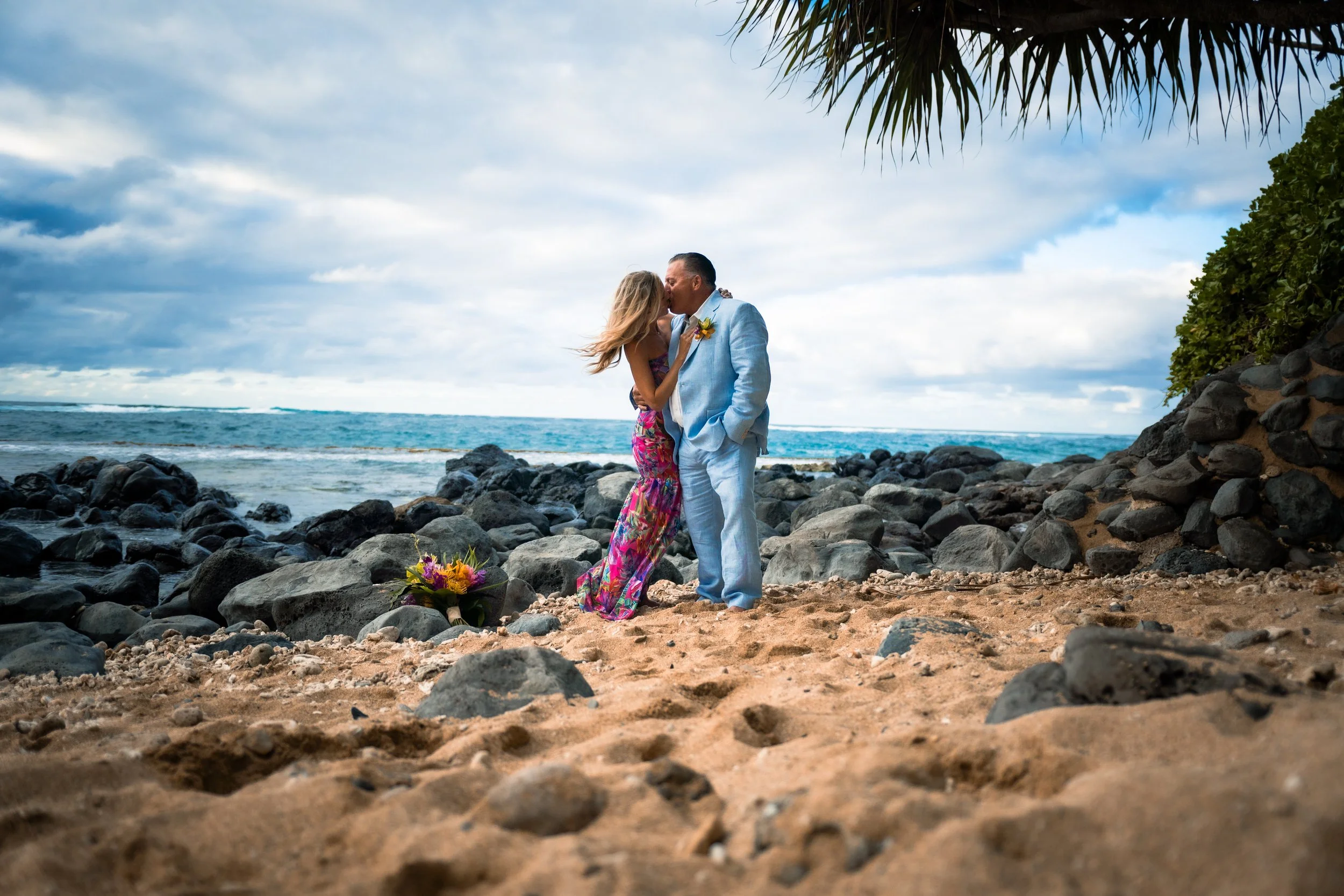 A couple dressed in formal attire sharing a kiss on a rocky beach under a cloudy sky, with a bouquet on the sand nearby.