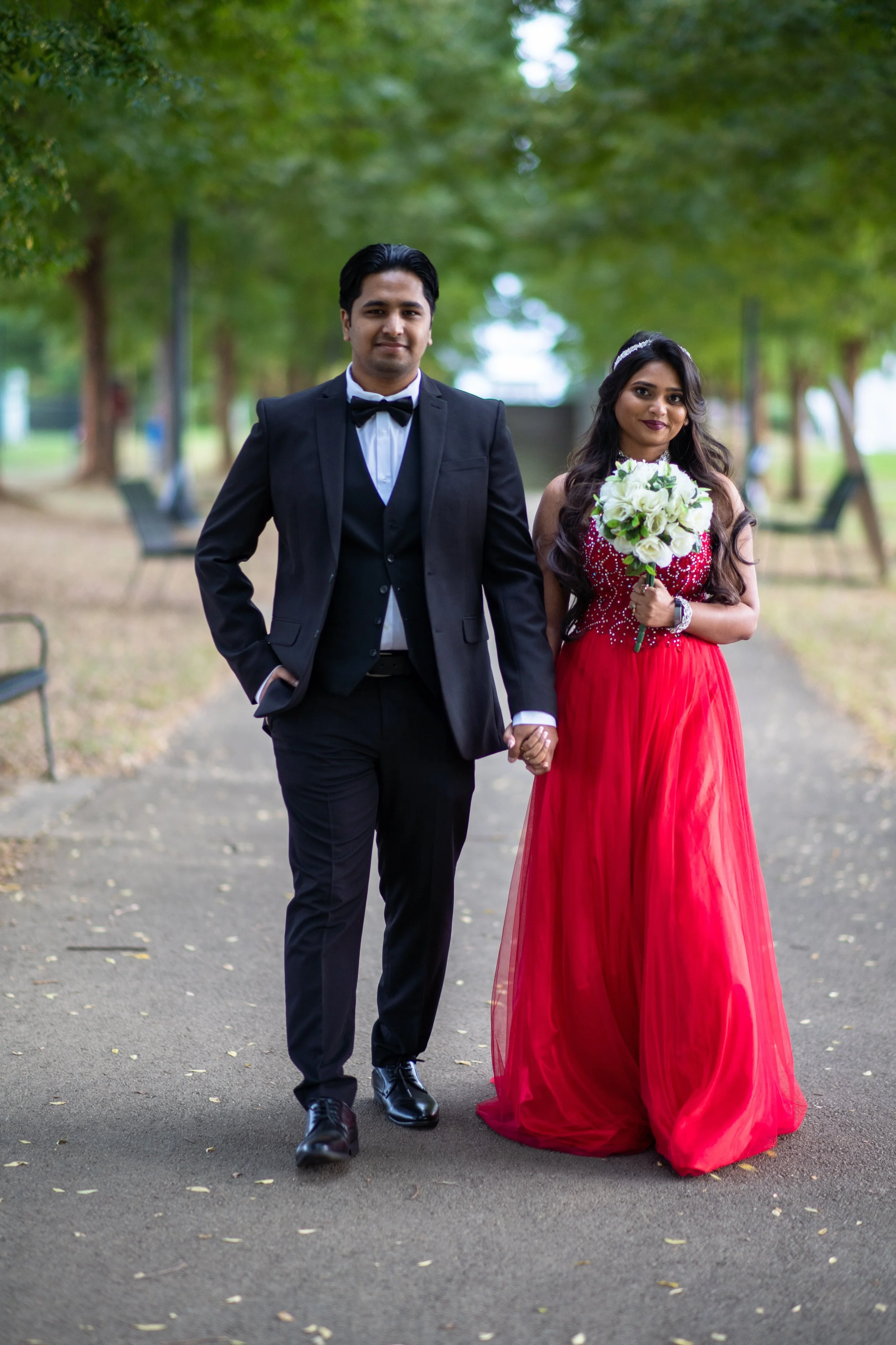 A couple dressed in formal attire holding hands and walking in a park with green trees around. The man is wearing a black tuxedo with a white shirt and black bow tie, and the woman is wearing a red gown and holding a bouquet of white flowers.