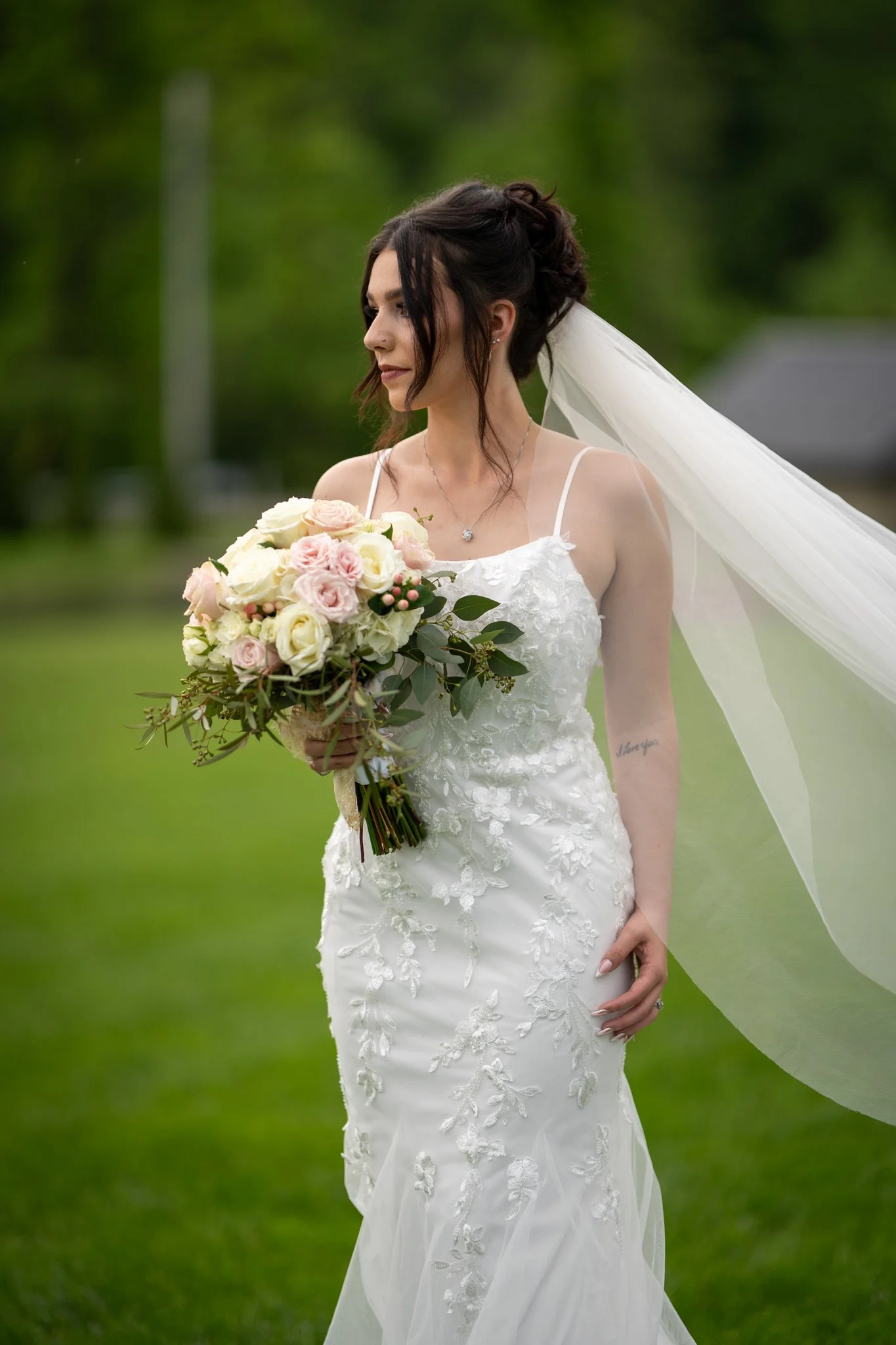 A bride in a white lace wedding dress holding a bouquet of roses and greenery outdoors.
