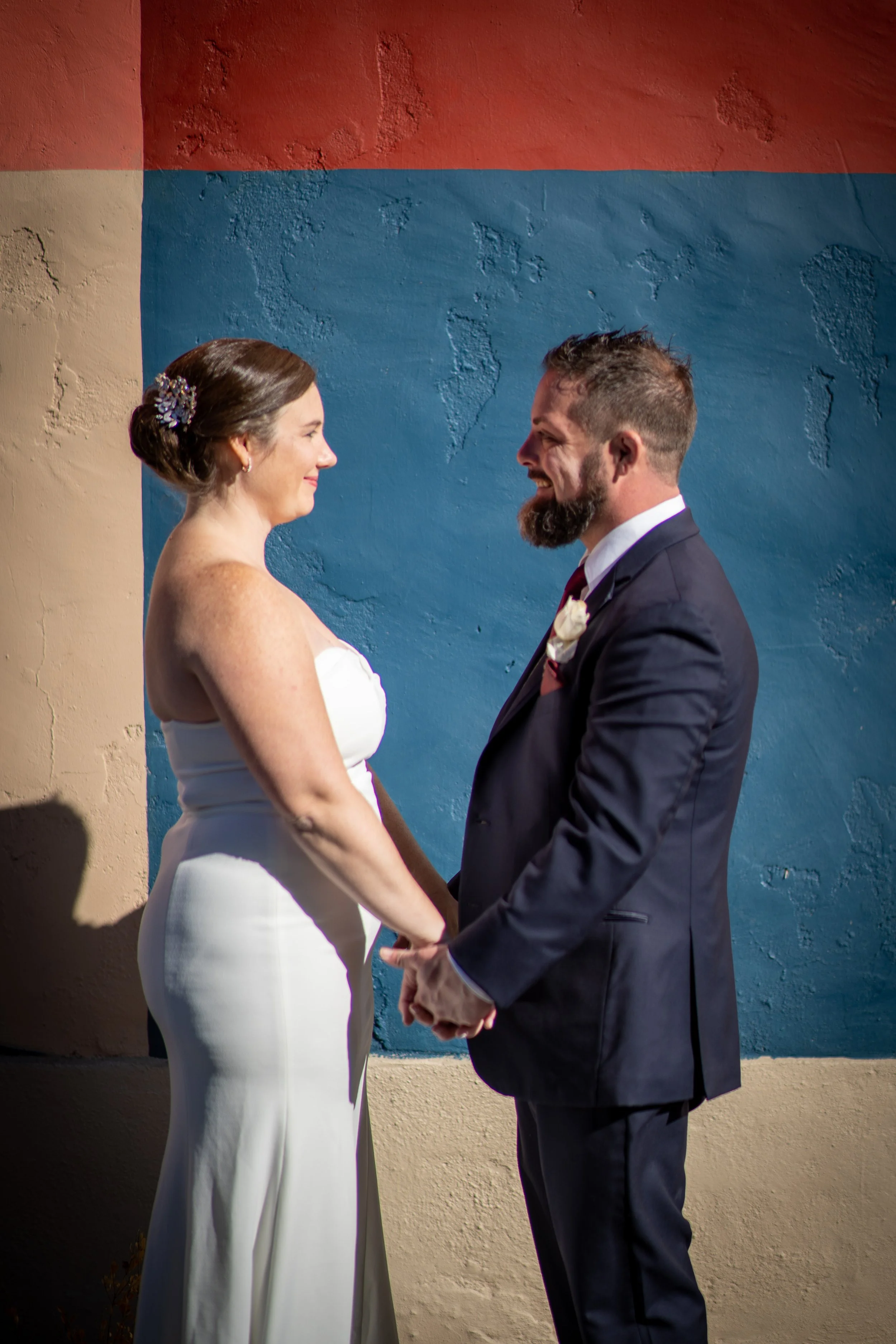 A couple getting married, facing each other and holding hands, with a painted colorful wall in the background.