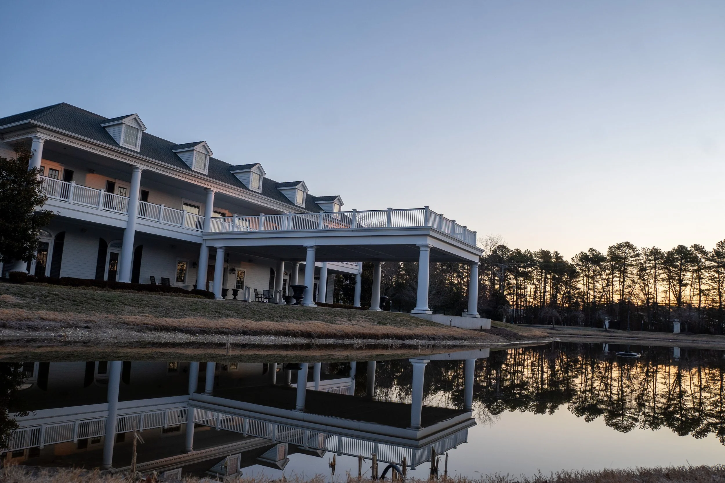 A large white multi-story house with a wraparound porch and multiple dormer windows, situated near a still body of water at sunset with trees in the background.