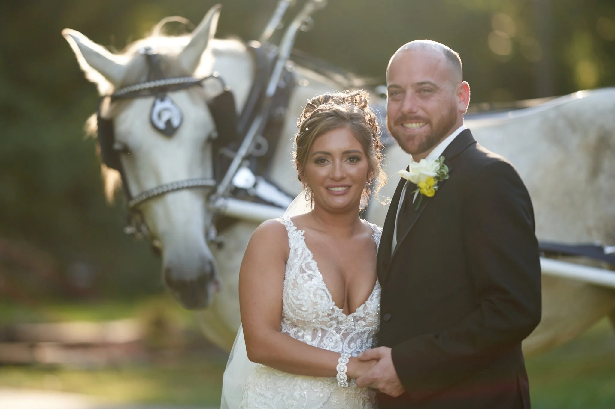Bride and groom smiling together outdoors near a white horse in a wedding photo.