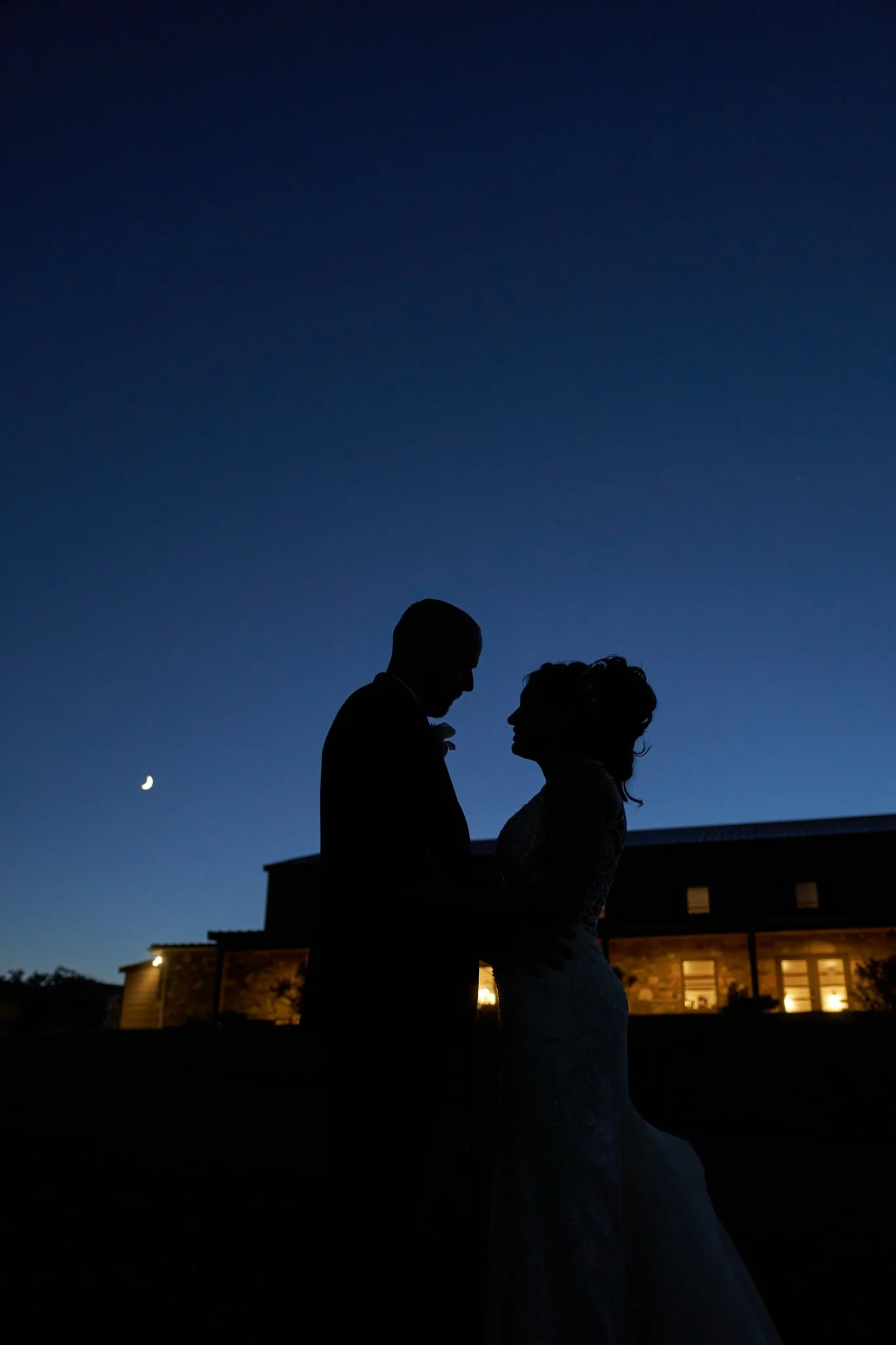 Silhouette of a bride and groom standing closely together outside during dusk, with a crescent moon in the sky and a building with illuminated windows in the background.